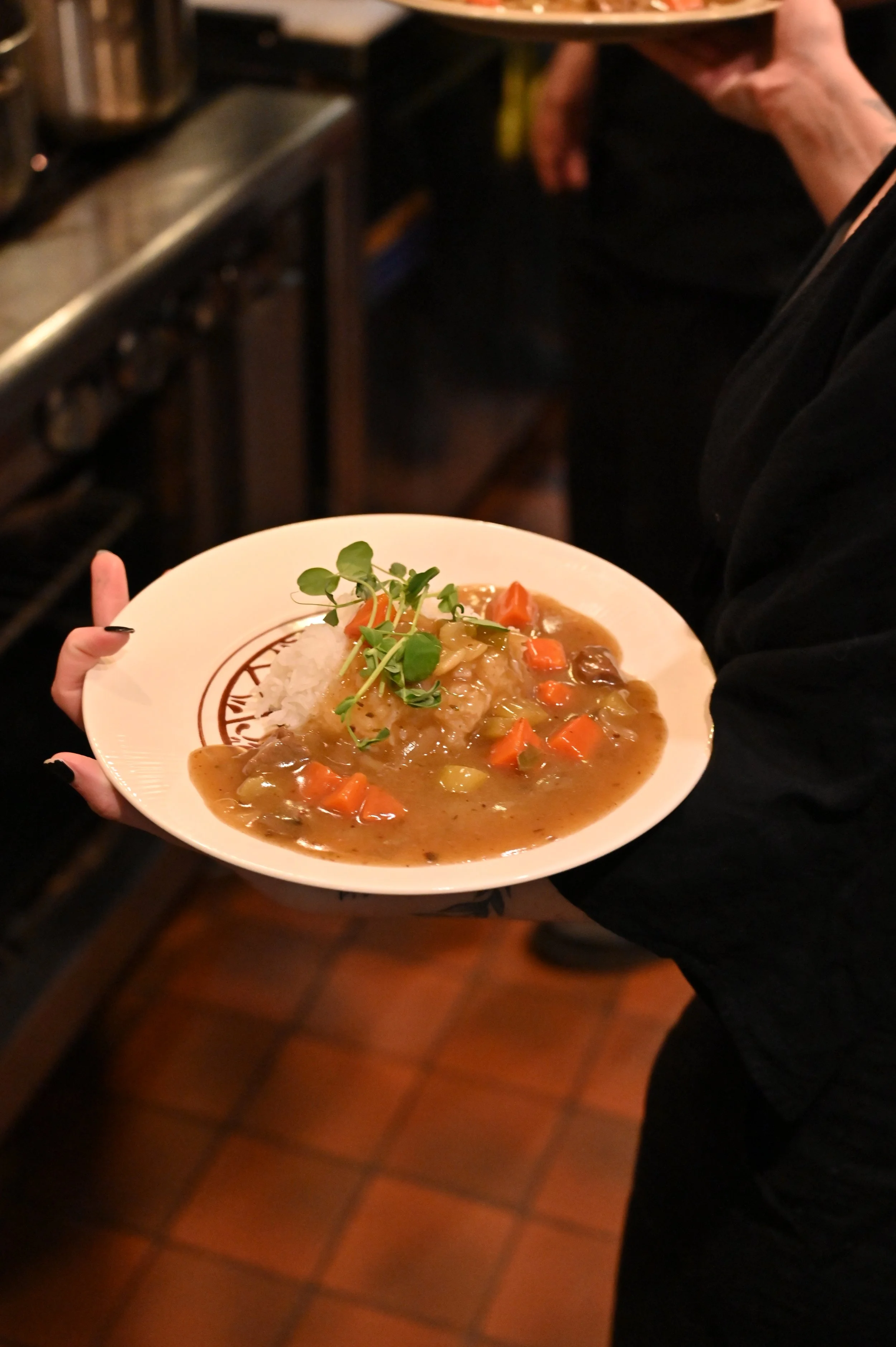 A person holding a plate of Japanese curry with rice and garnished with microgreens, in a restaurant setting.
