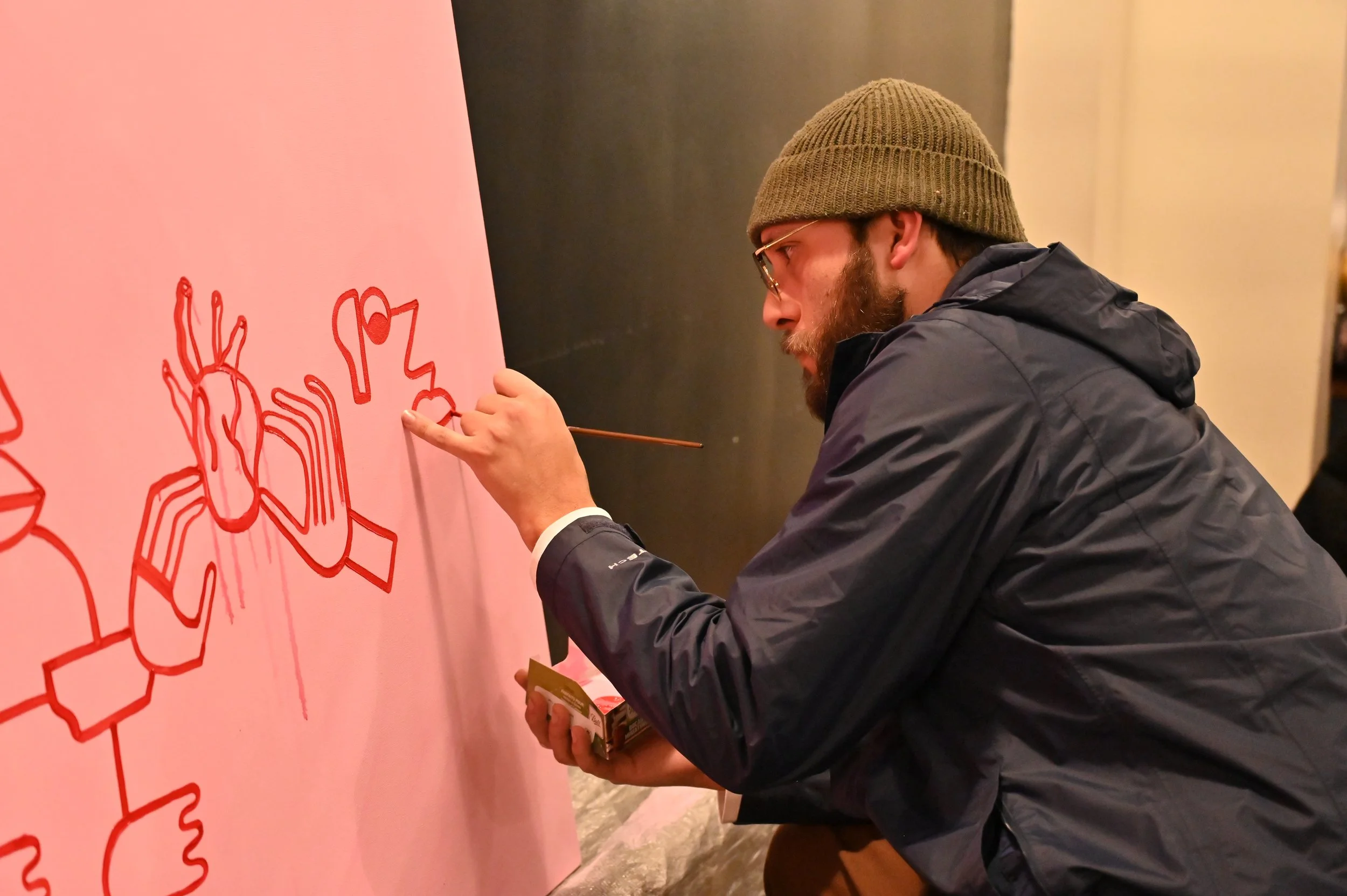 A man with a beanie hat, glasses, and a dark jacket is drawing with a red marker on a pink canvas.