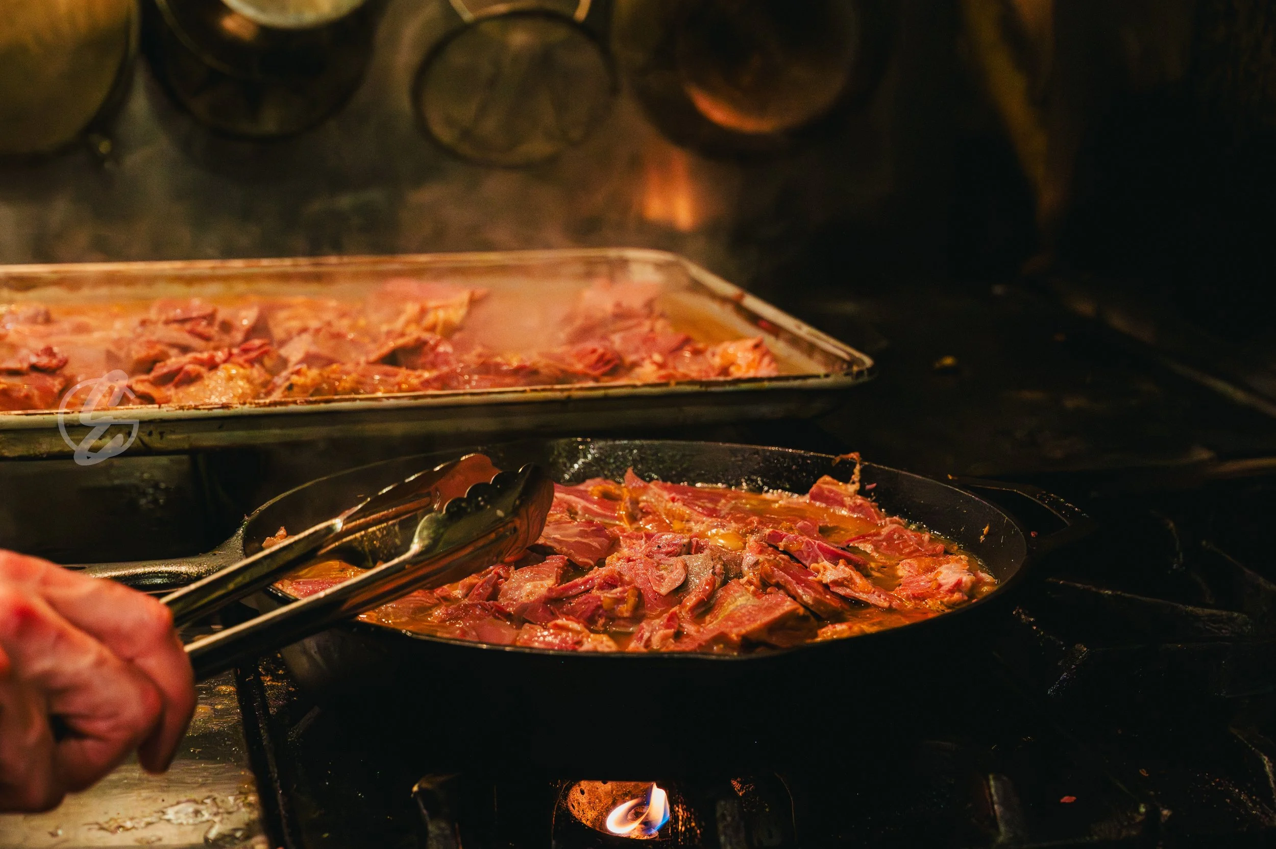 Cooking meat on a stove with tongs in a kitchen, with a baking tray of raw meat in the background.