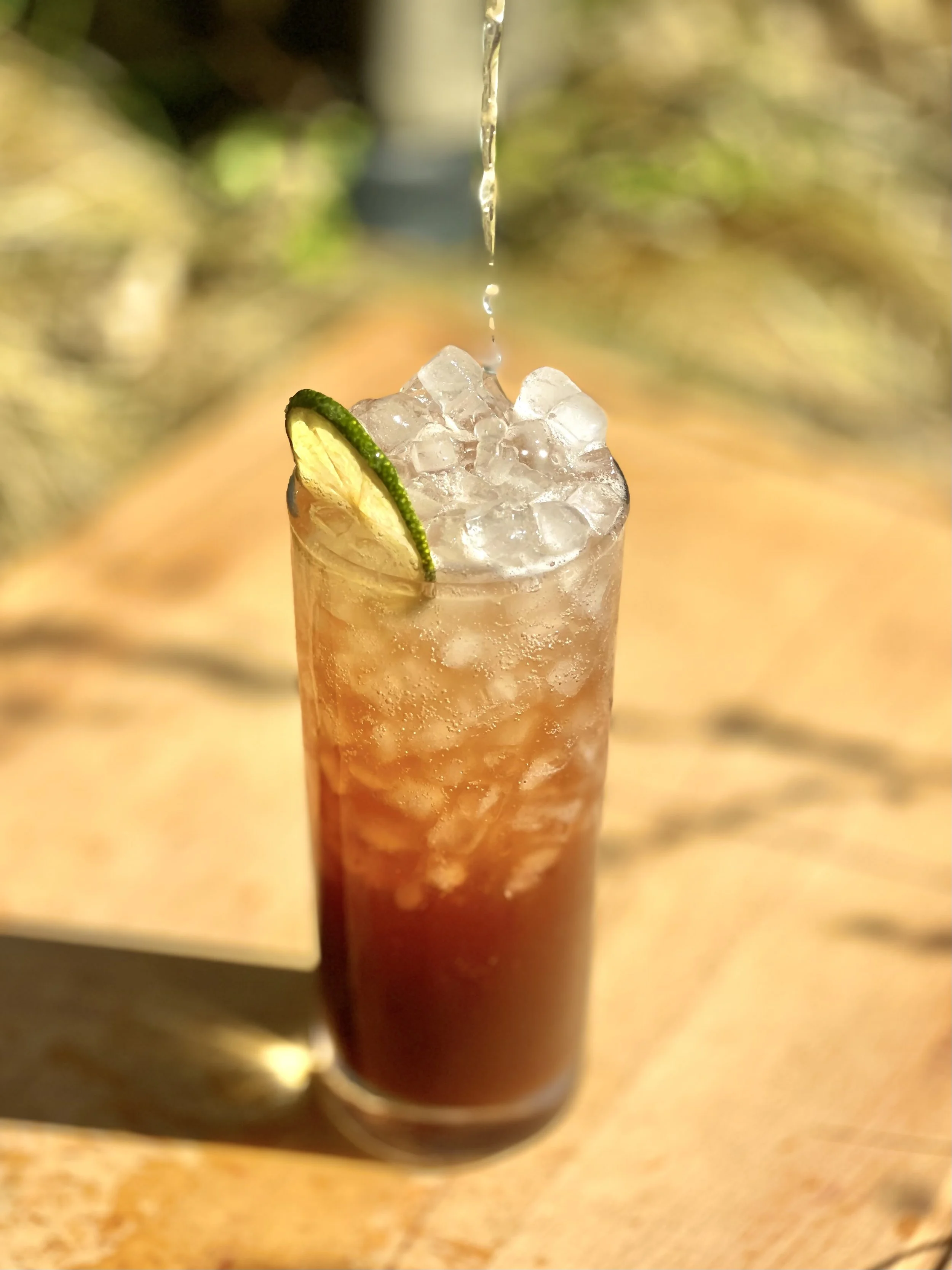 A tall glass of iced tea with condensation, ice cubes, a lime wedge garnishing the rim, and water being poured on top, set on an outdoor surface with blurred greenery in the background.
