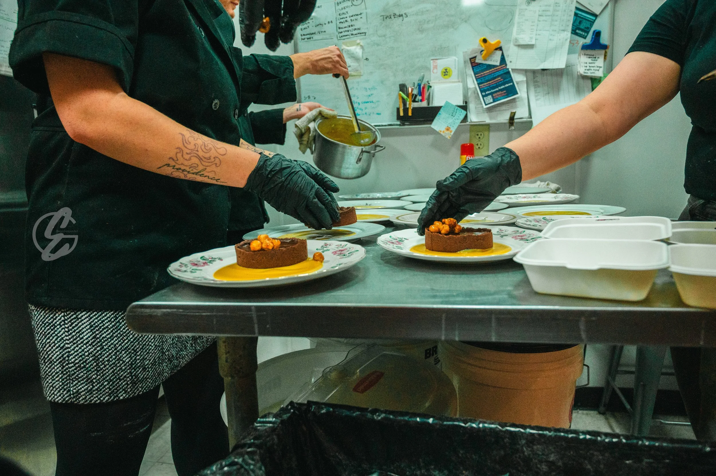 Chefs are preparing plated desserts in a commercial kitchen, placing orange garnishes on chocolate dessert discs with a yellow sauce underneath.