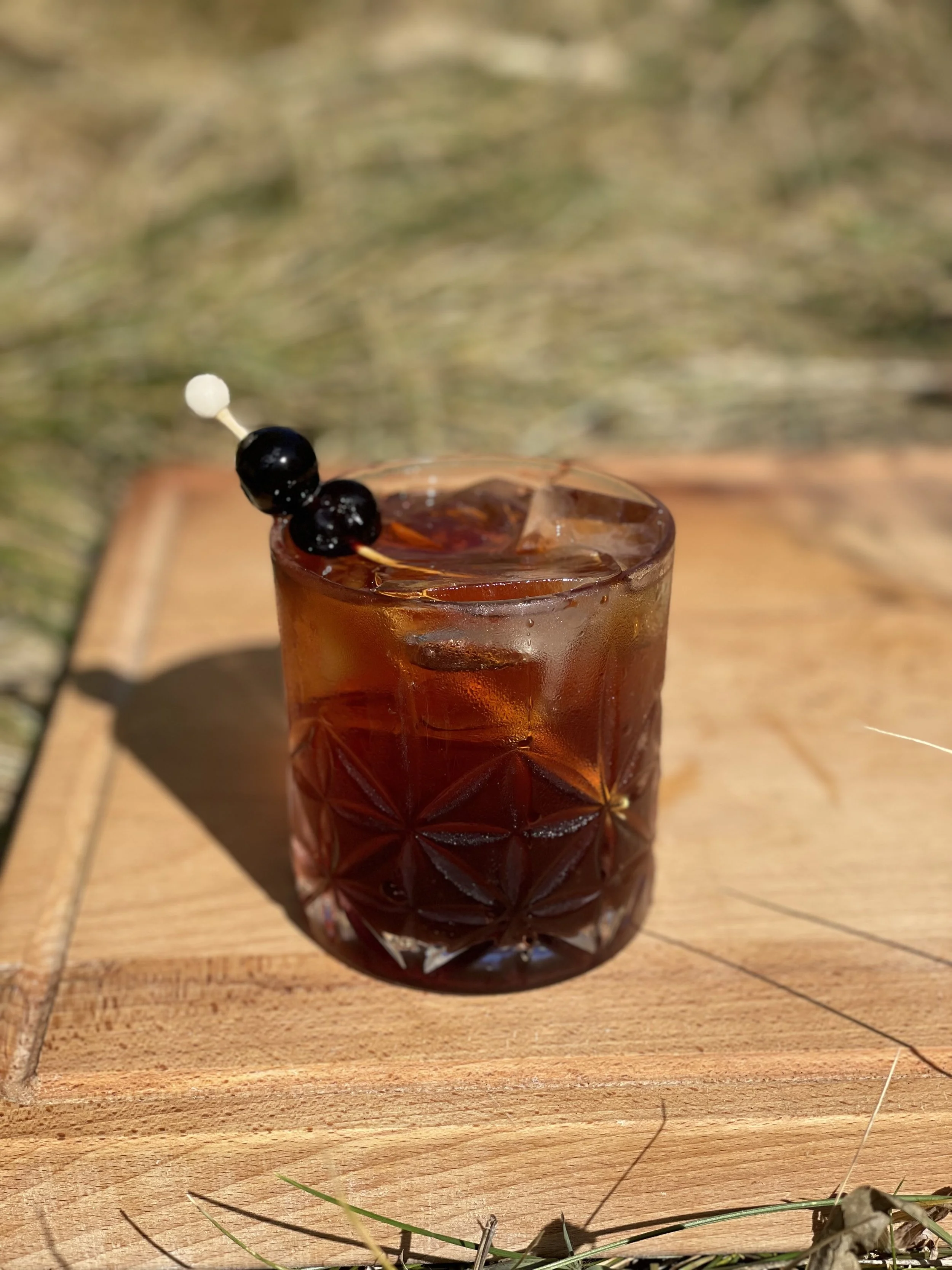 A glass of dark cocktail with two black berries and a white berry on a toothpick, placed on a wooden surface outdoors.