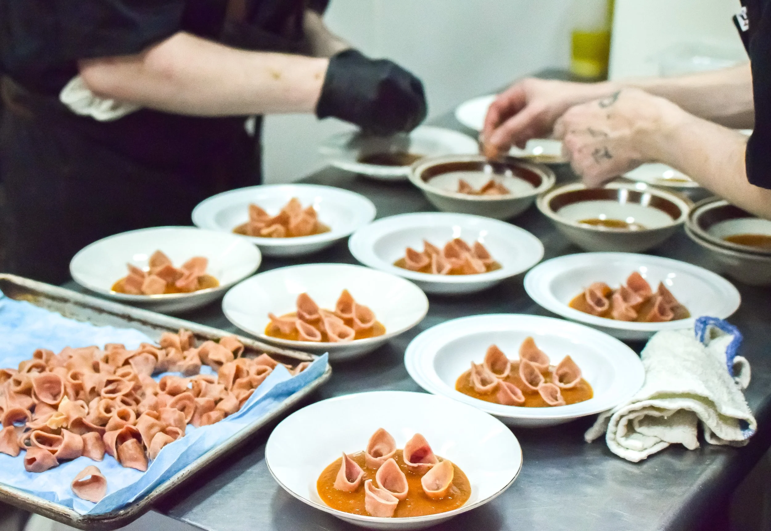 Food preparation scene with plates of uncooked tortellini on a tray and a table of bowls filled with are filled with sauce, as two people work together, one wearing black gloves.