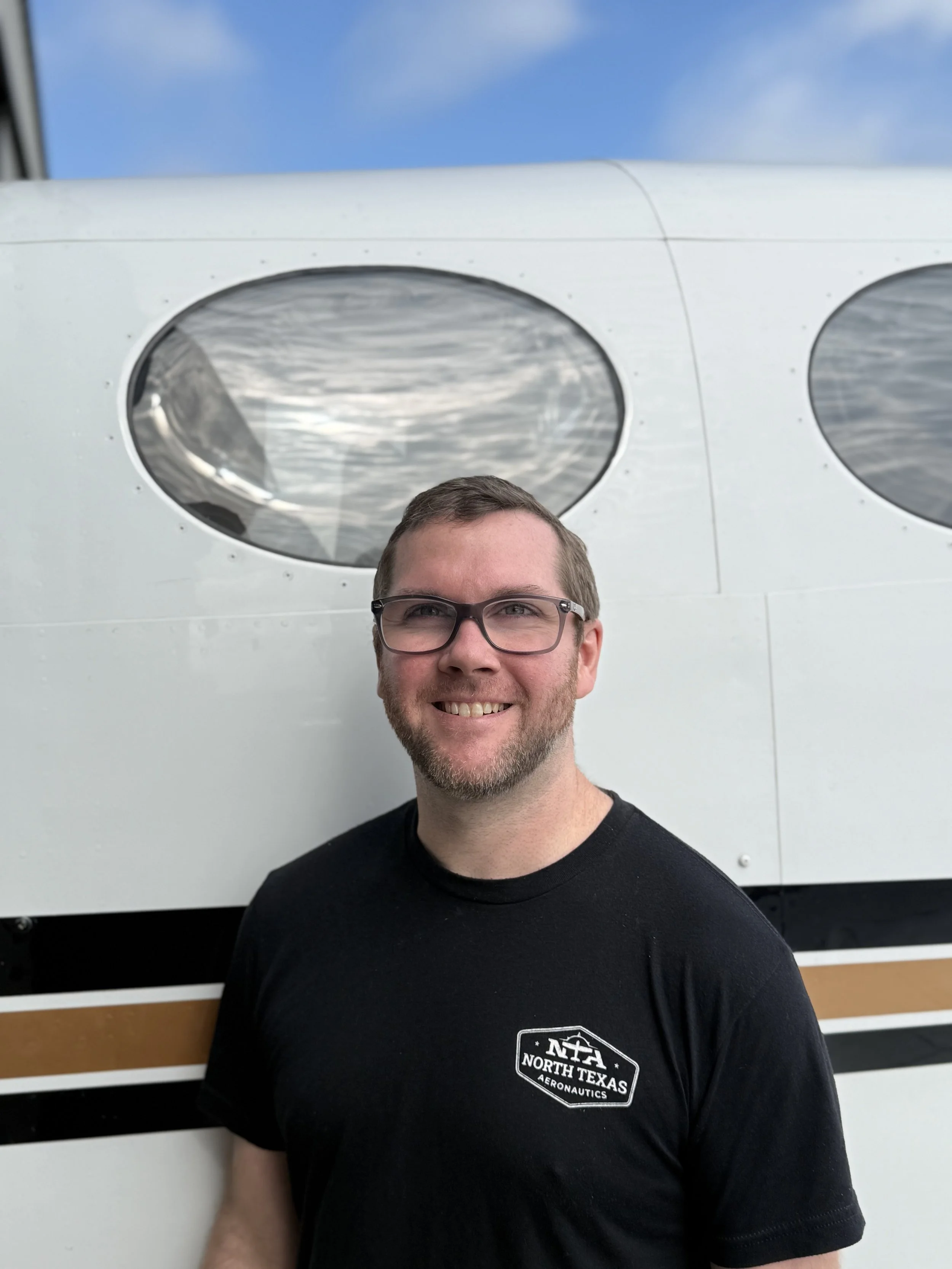 A smiling man with glasses and a beard standing in front of a vintage aircraft inside a hangar.