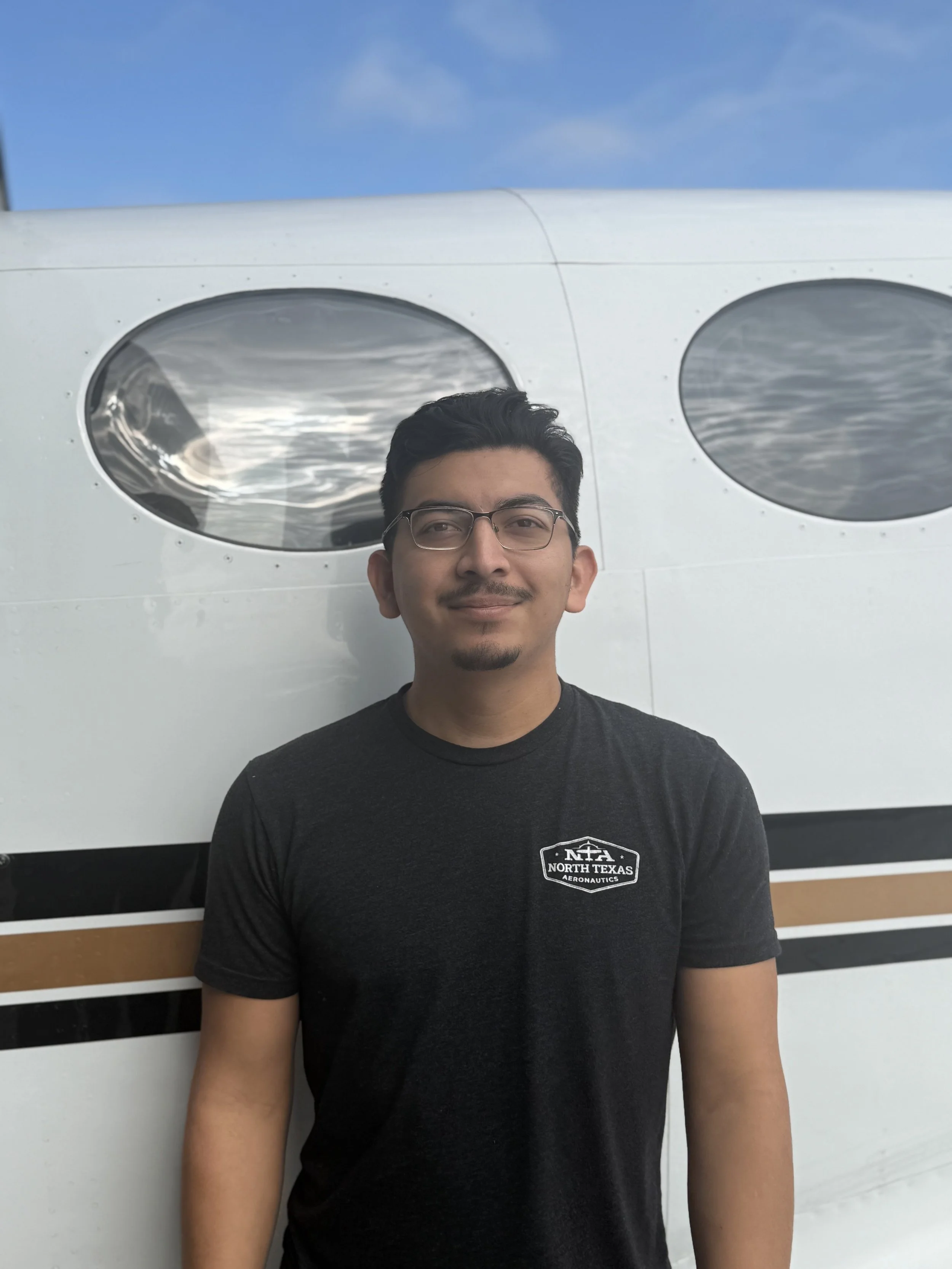 A man with glasses, a goatee, and black hair, smiling, standing in front of an aircraft inside a hangar.