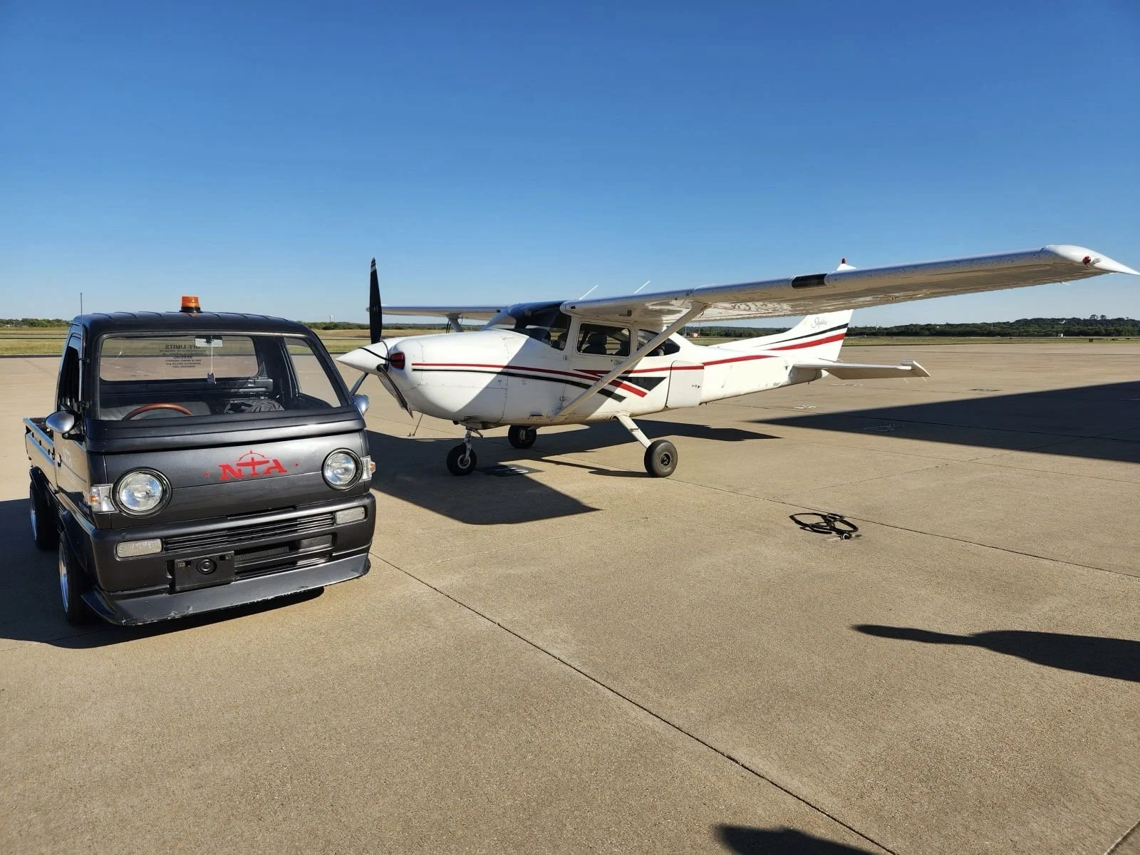 Small black utility vehicle with red letters 'NTA' on the front, parked on a tarmac next to a white single-engine propeller airplane with red and black stripes, under a clear blue sky.