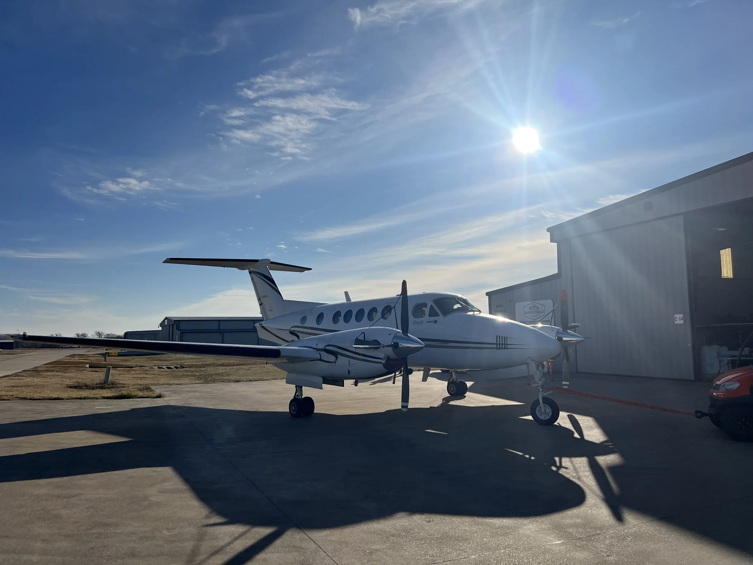 Small private jet parked on tarmac near hangar under a clear, sunny sky with some wispy clouds.