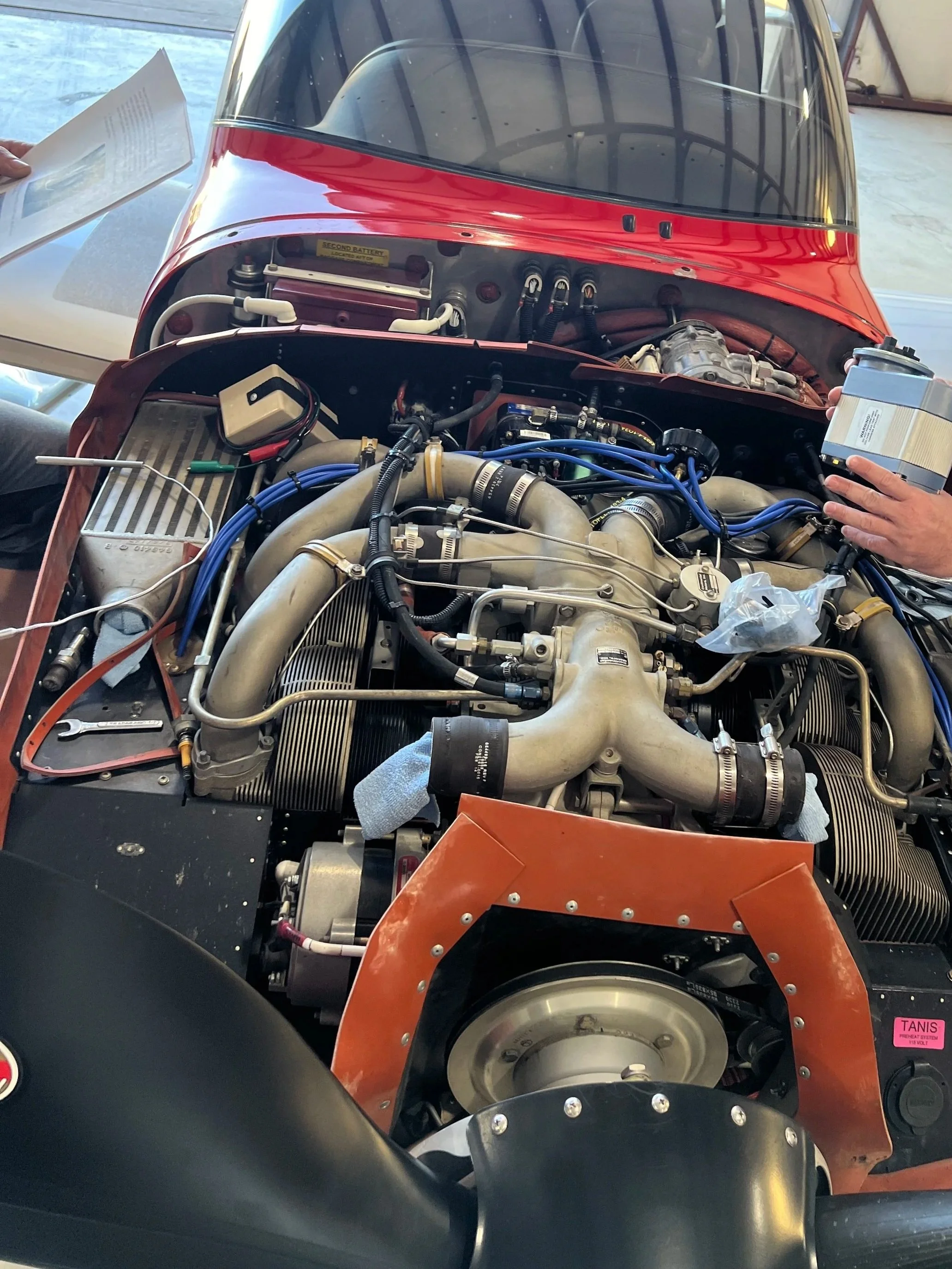 Close-up of a car engine inside its red and black body, with various hoses, wires, and mechanical components visible. Hands are seen holding a container and document near the engine.