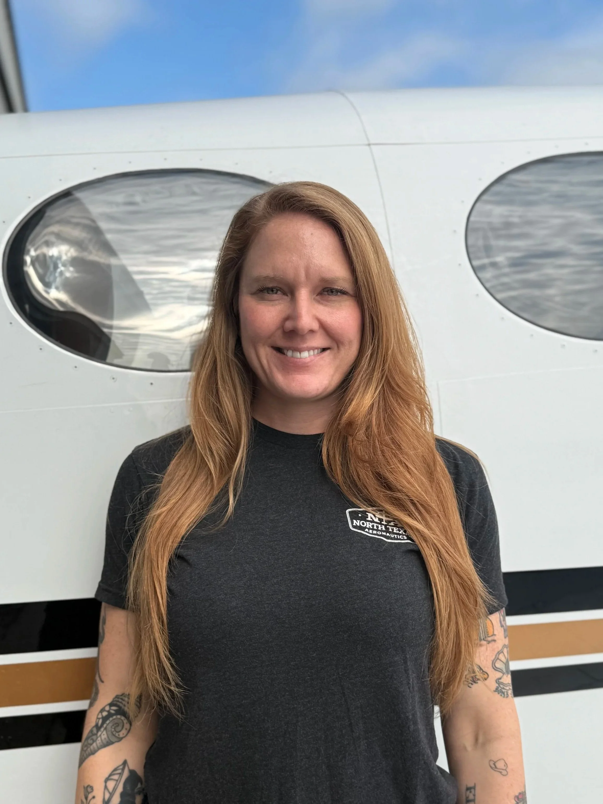 A woman with long, wavy red hair and multiple tattoos on her arms, smiling in front of an aircraft in an indoor hangar.