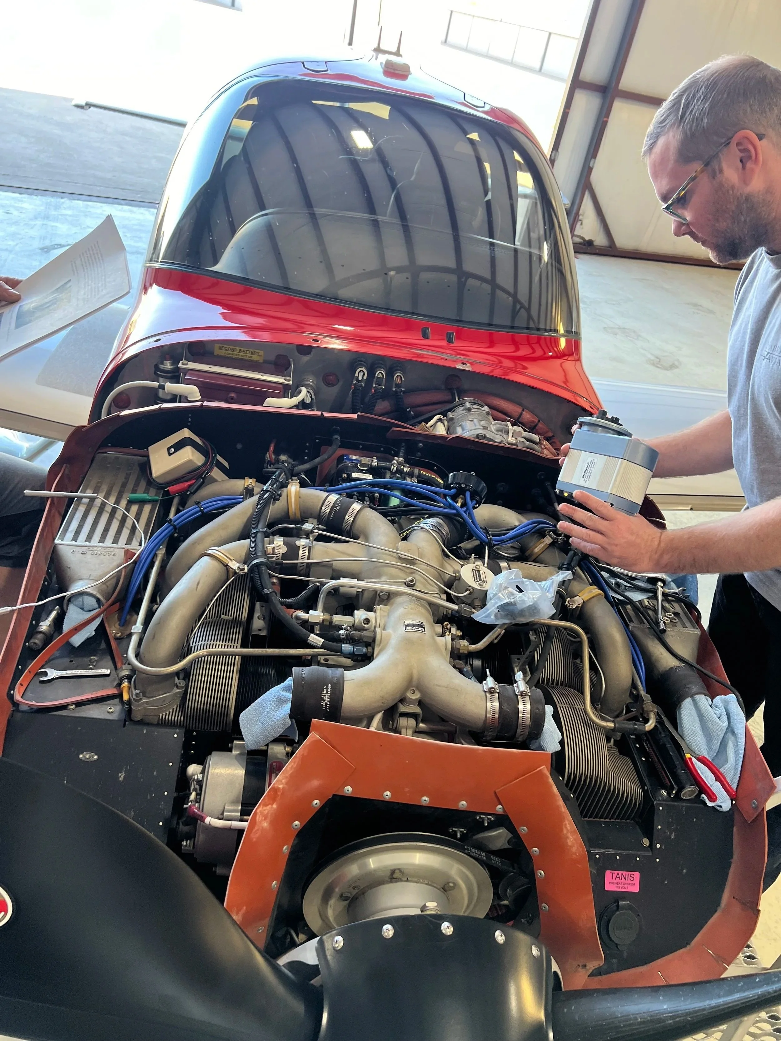 A person with short hair and glasses working on the engine of a small red aircraft inside a hangar. The aircraft's canopy is transparent, revealing the cockpit.