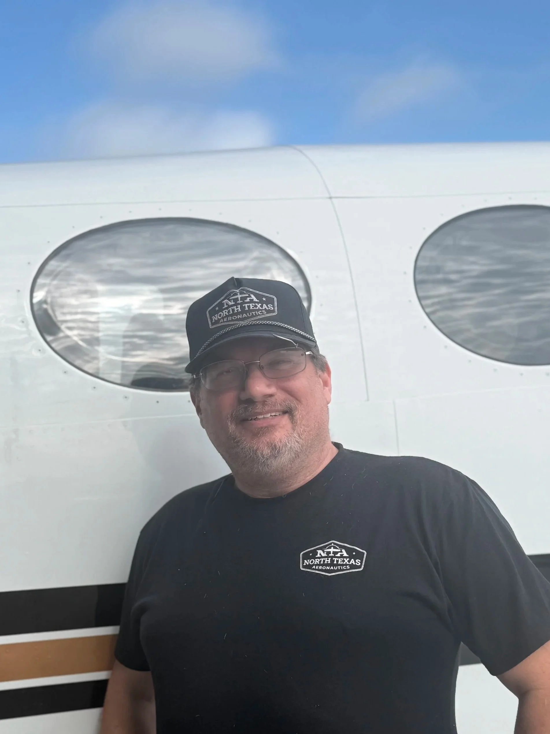A man with glasses and a beard, wearing a Carhartt jacket and a North Texas Aeronautics cap, is smiling and touching his chest with his right hand, standing in front of an aircraft.