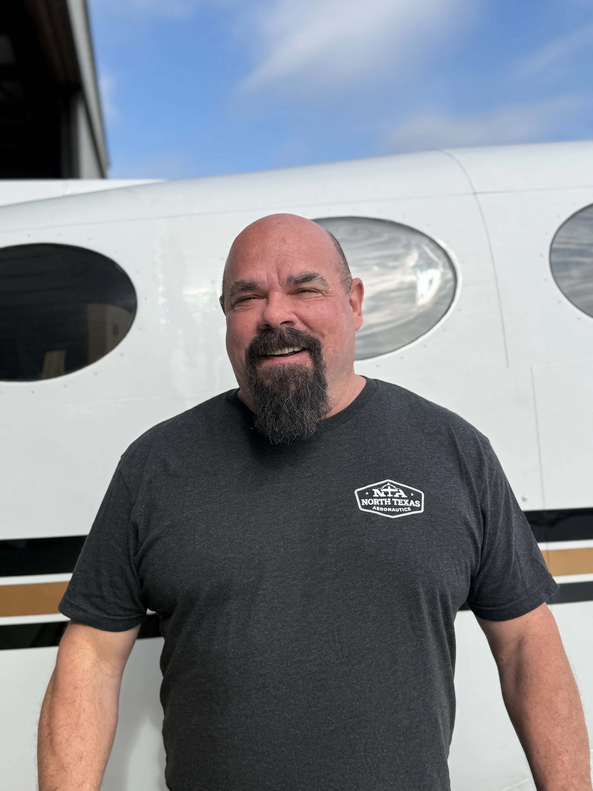 A man with a shaved head and dark beard smiling in front of a small airplane in a hangar.