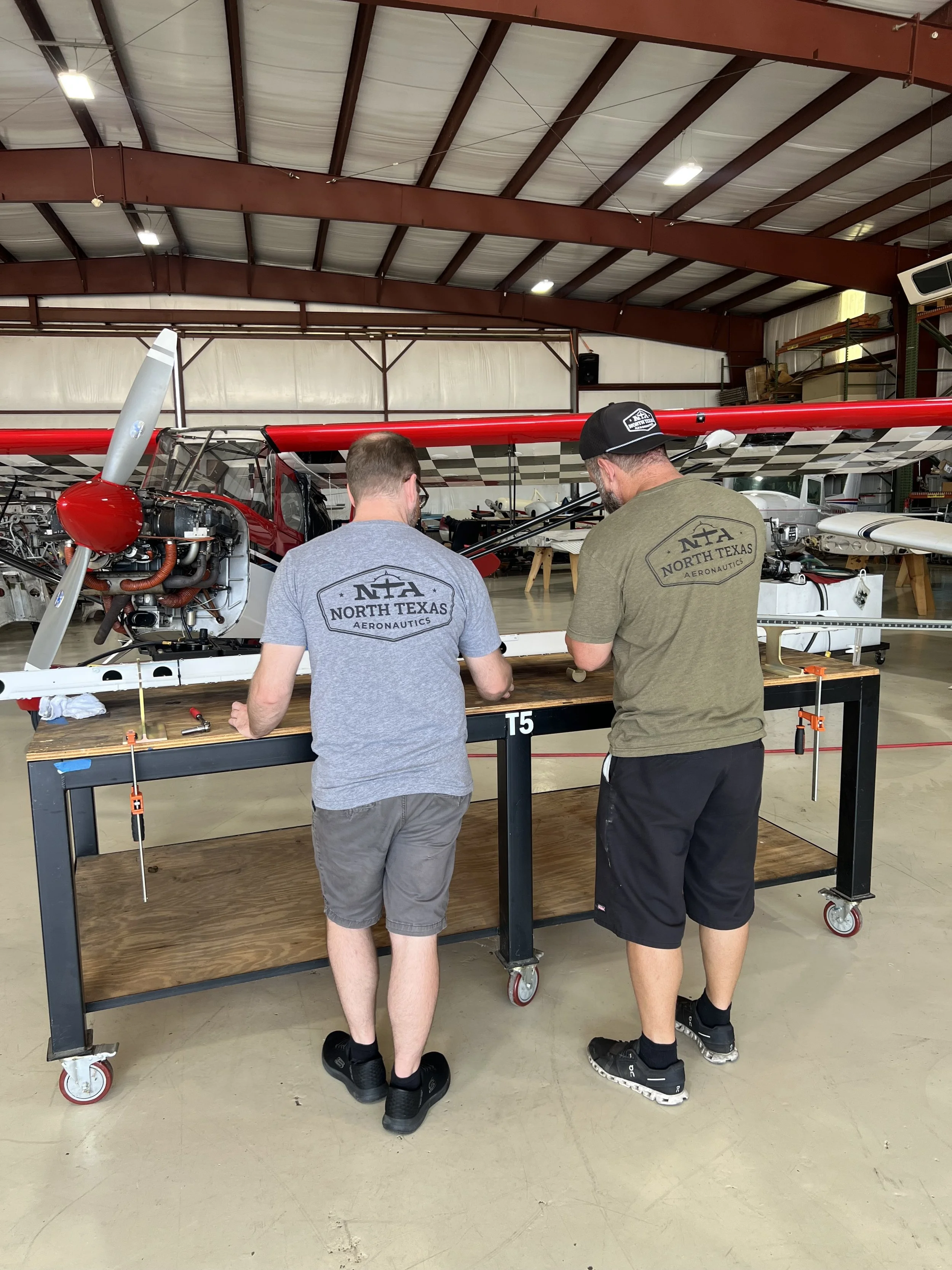 Two men stand at a workbench inspecting a small aircraft in an aircraft hangar. The aircraft has a red wing with a black and white checkered pattern underneath. The hangar has a high ceiling with brown metal beams and various aircraft in the background.