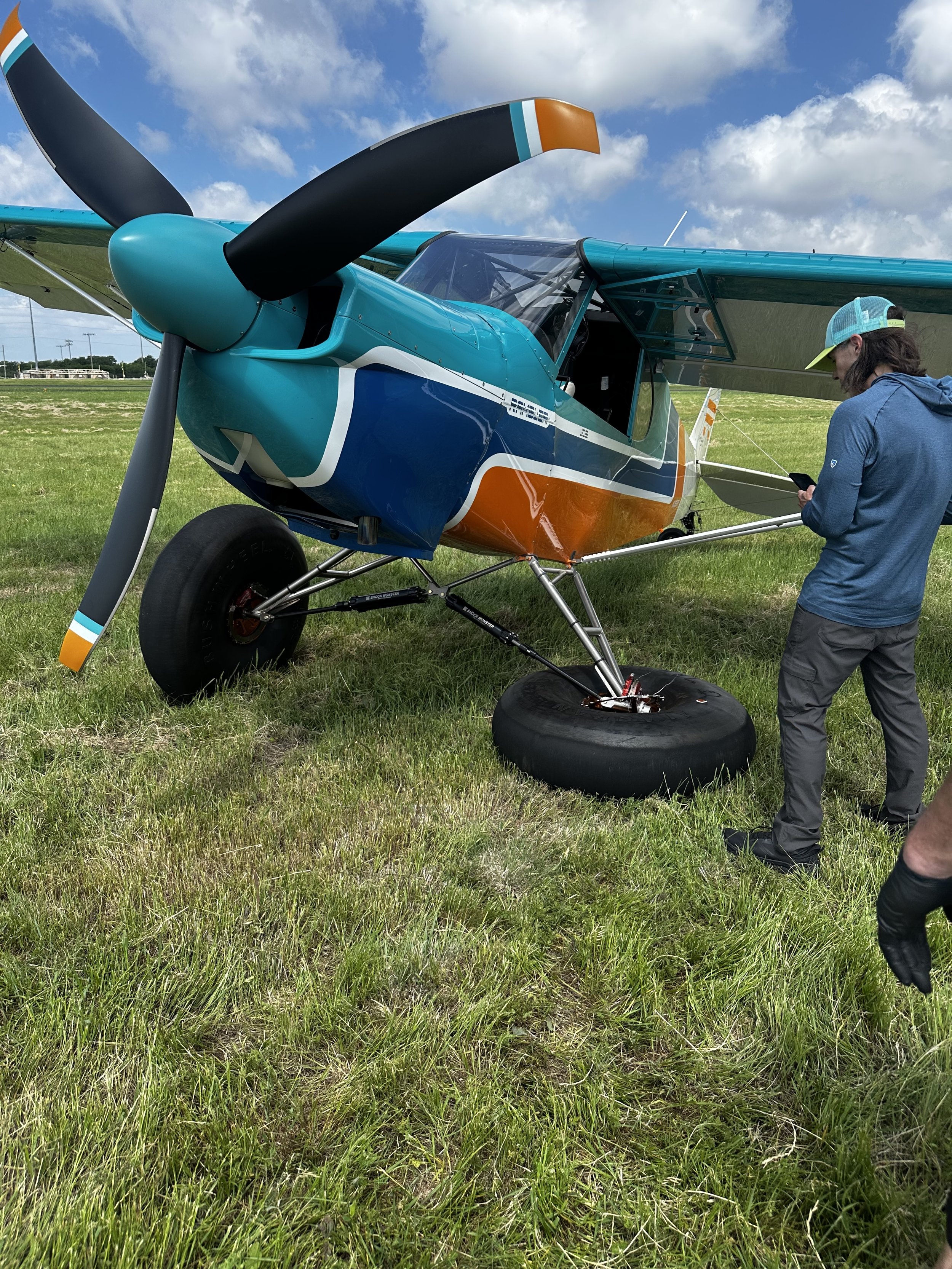 A small aircraft with a ground crew person standing beside it, looking at their phone, on a grassy field under a partly cloudy sky.