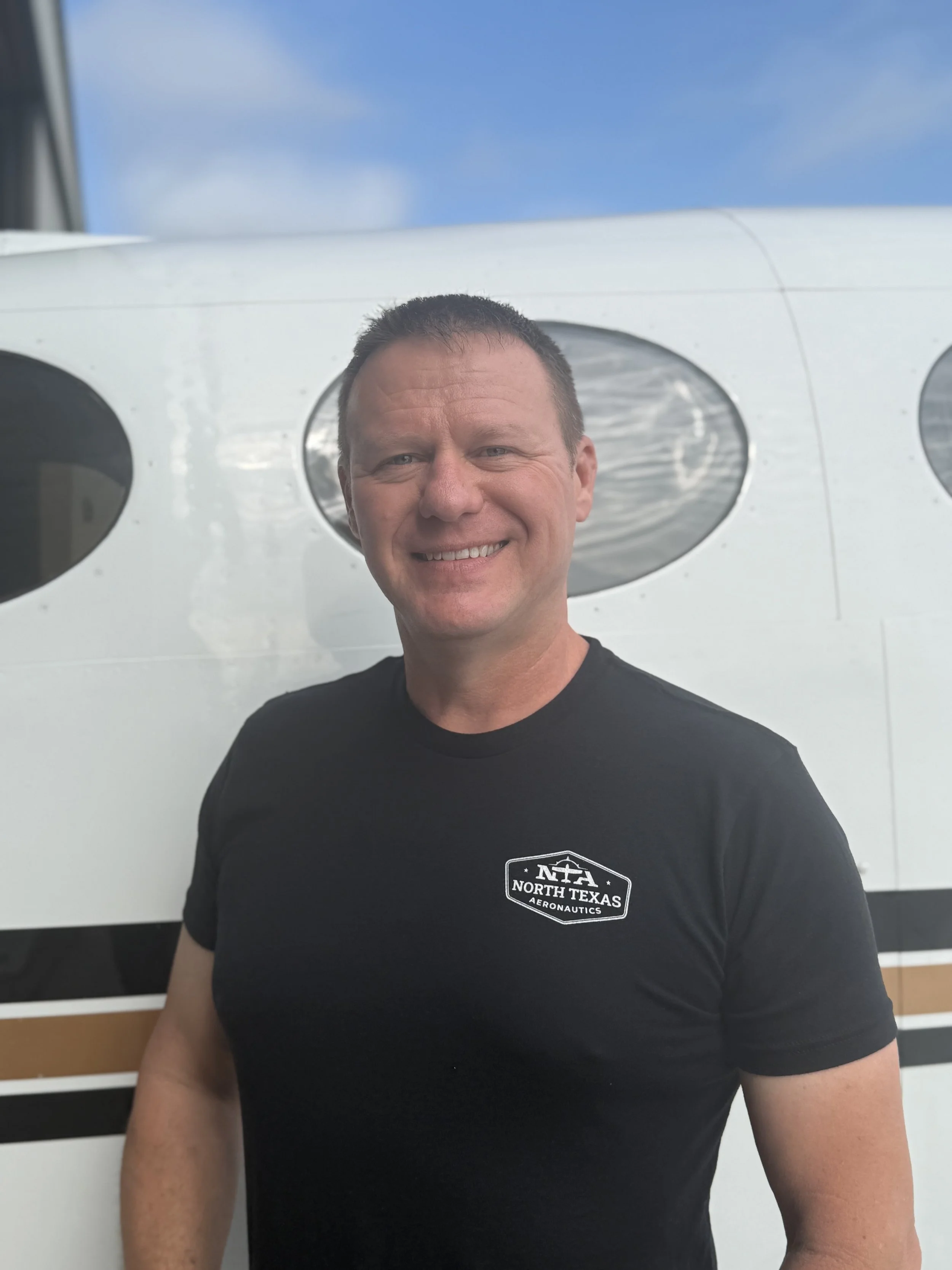 A smiling man wearing a gray t-shirt standing inside an aircraft hangar with small airplanes in the background.
