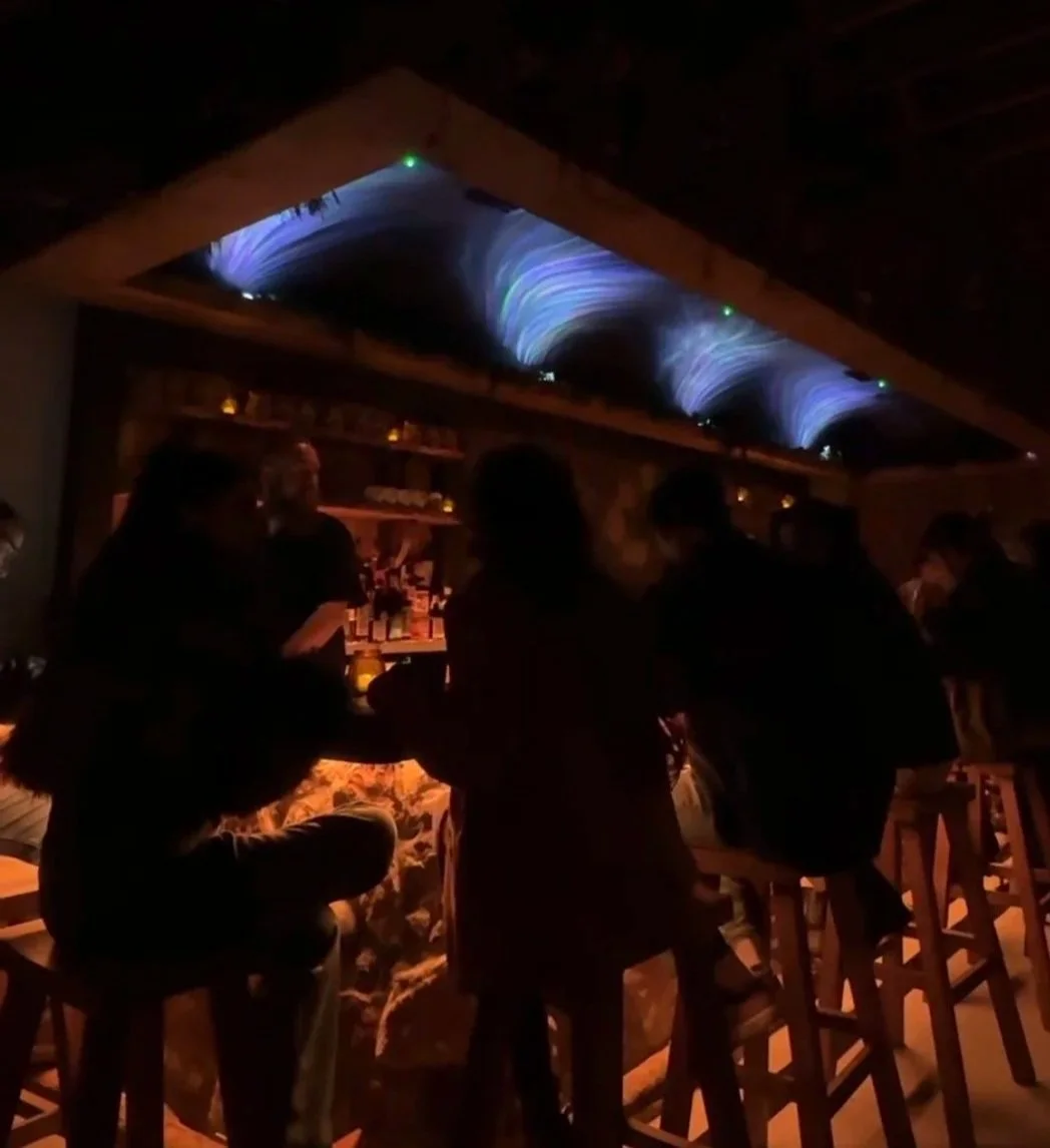 Silhouettes of people sitting and standing at a bar in a dimly lit restaurant or pub with an illuminated ceiling featuring blue and purple wave-like patterns.