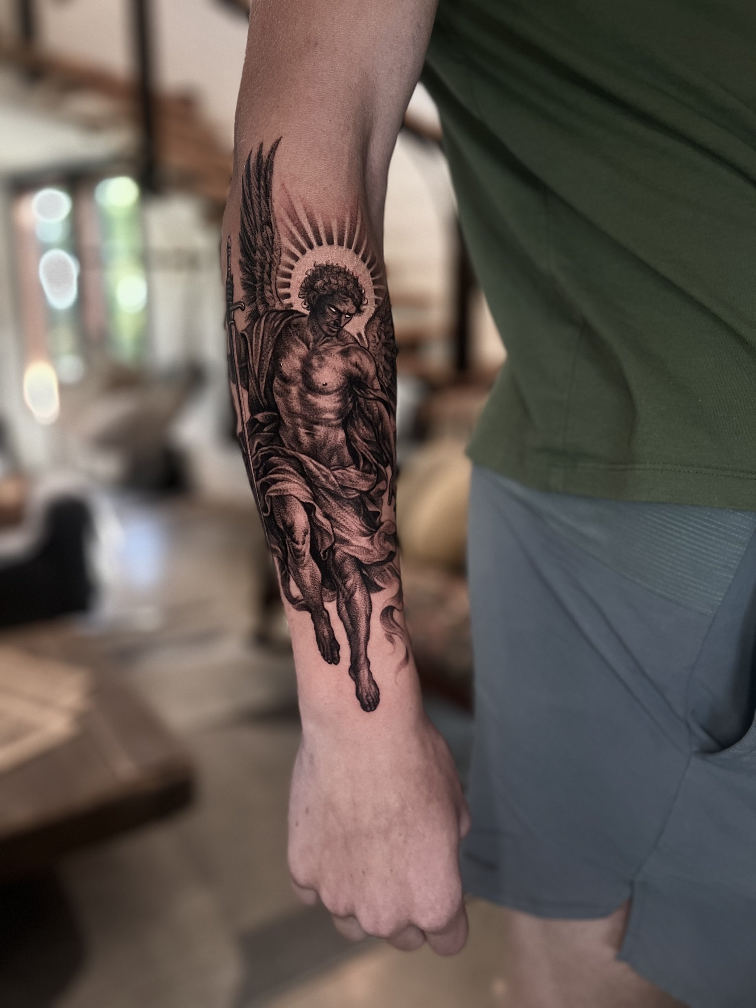 Tattoo of an angel or a biblical figure with wings and a halo, holding a sword, on a person's forearm.