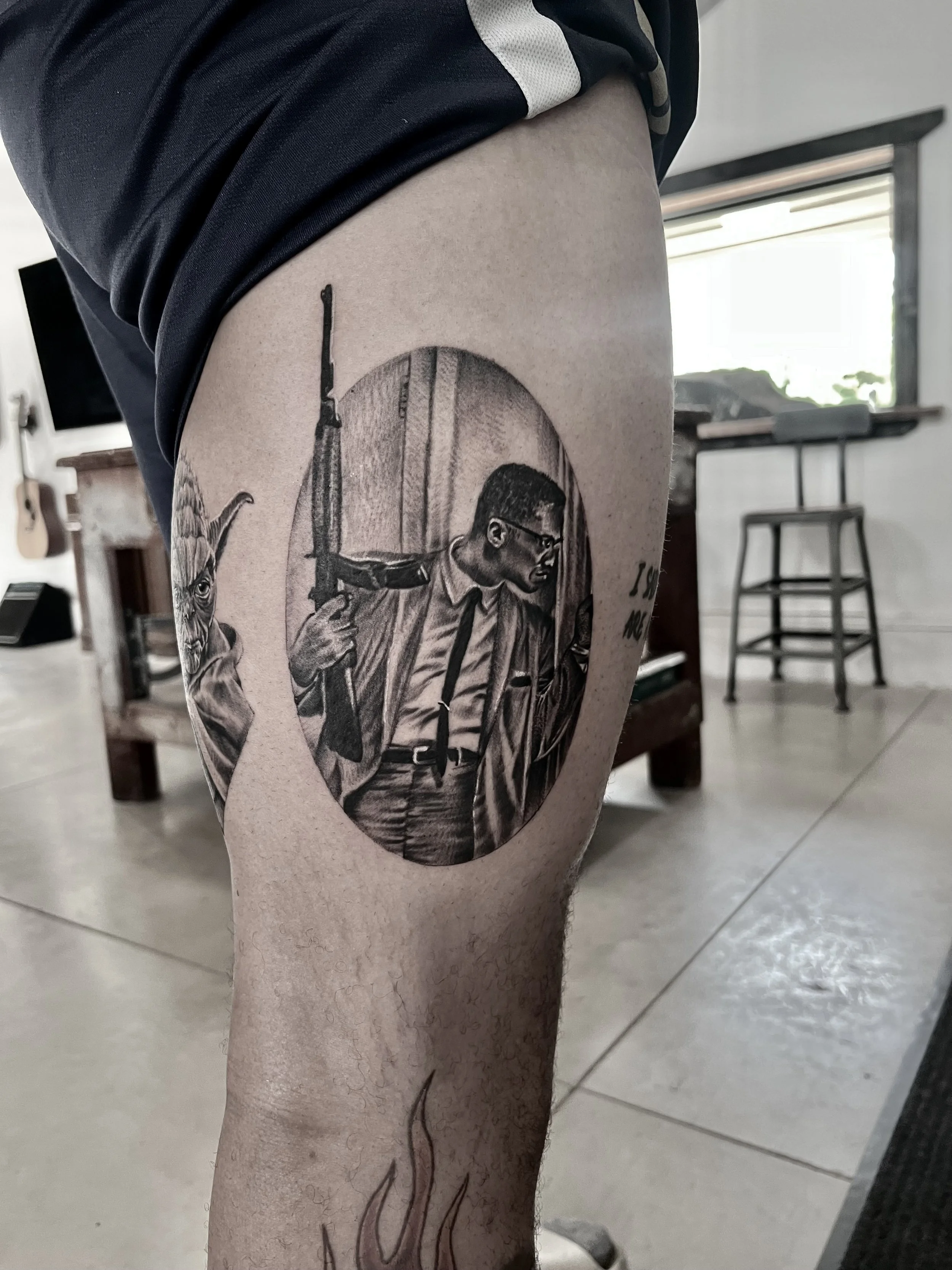 Tattoo of a man in a suit and glasses holding an assault rifle, with a creature resembling an alien on his shoulder, inside an oval frame on the thigh.