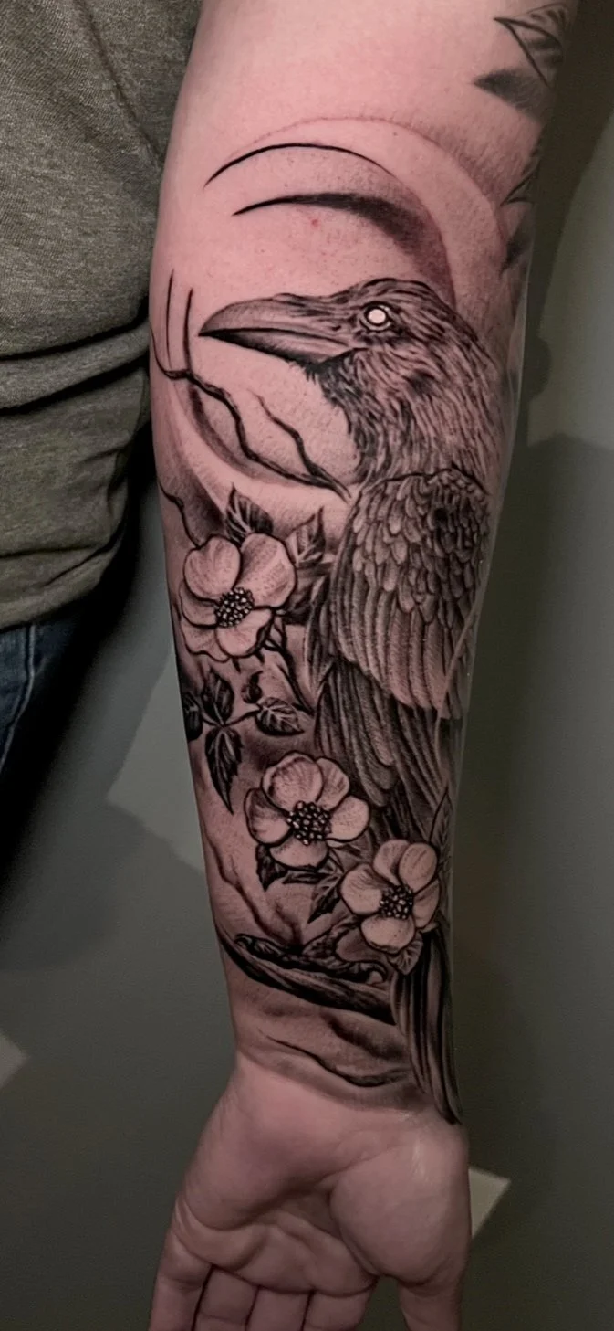 Black and gray tattoo of a bird with detailed feathers, perched on a branch with blooming flowers, on a person's forearm.