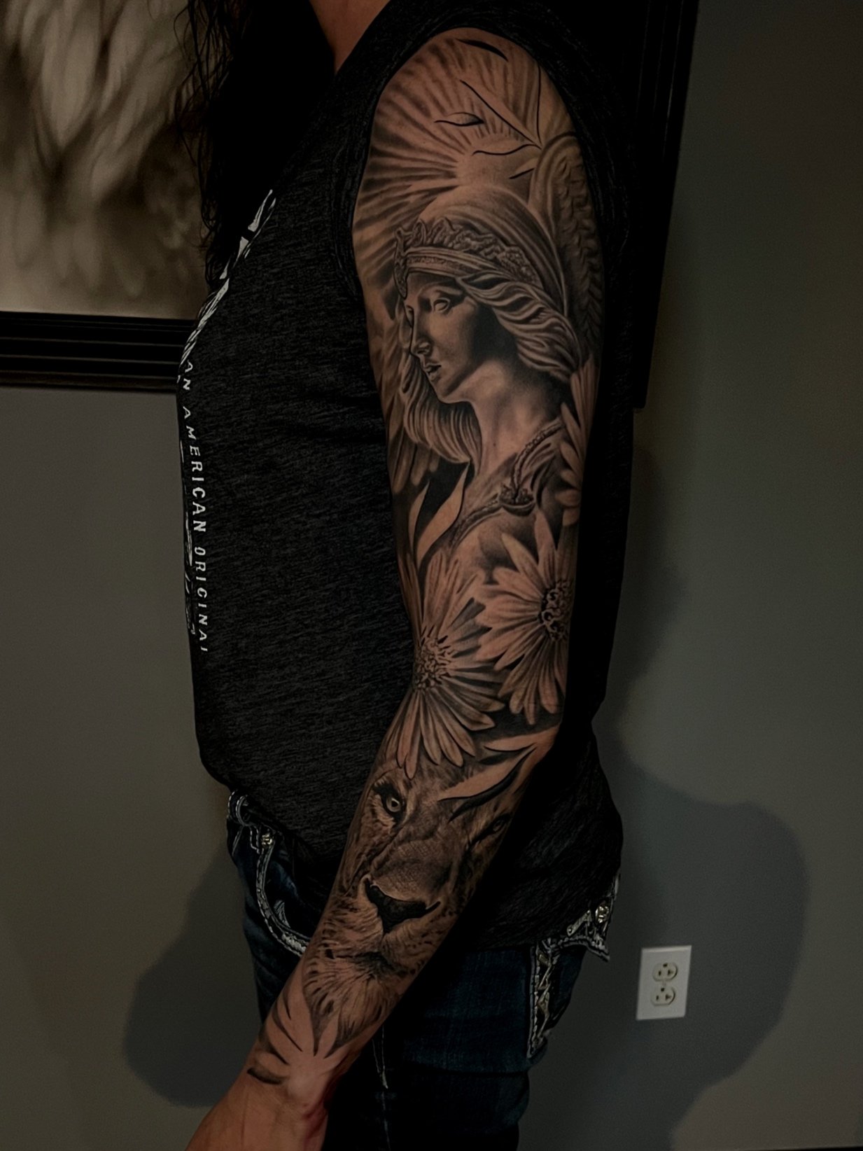 A person with a detailed black and gray tattoo sleeve featuring a woman's face with long hair and a feathered headdress, surrounded by large flowers and a lion's face on the forearm.