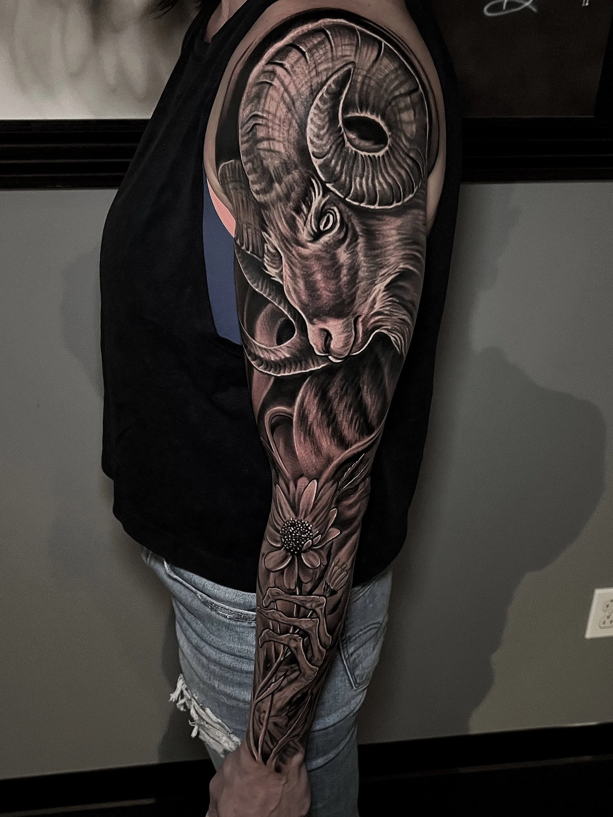 A detailed black and gray tattoo of a ram's head with large, curled horns, a realistic face, and a flower with long petals extending down the arm, with the person wearing a black shirt and ripped jeans.
