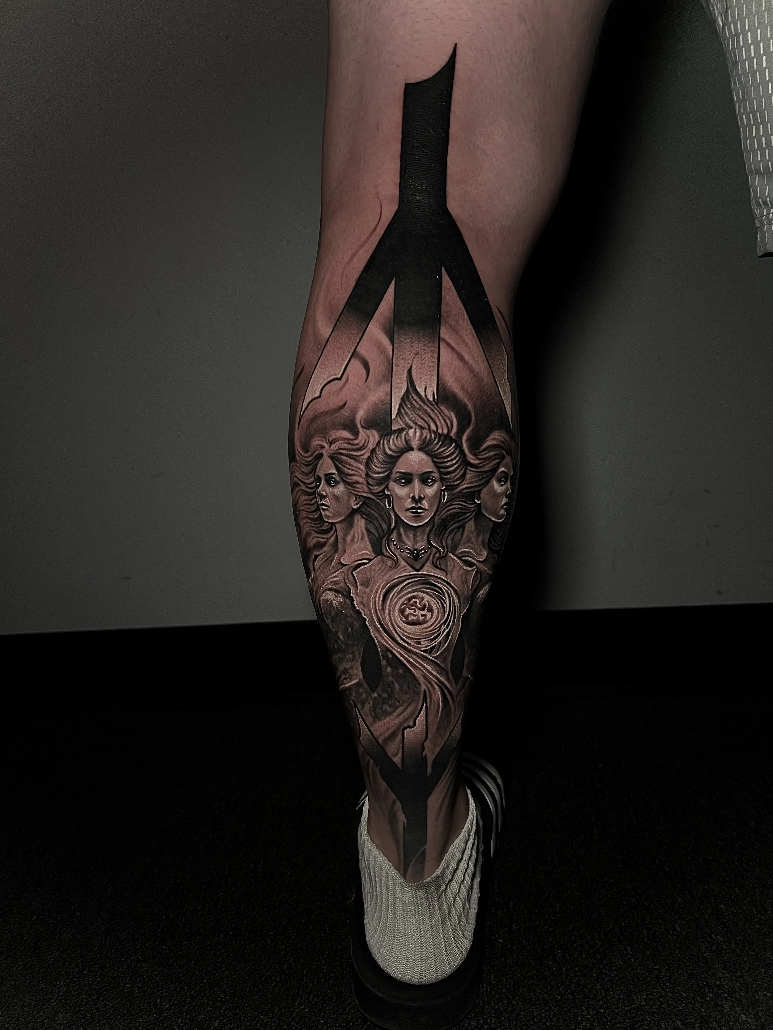 Tattoo of three women with flowing hair, with a large, bold, black geometric design above and around them.