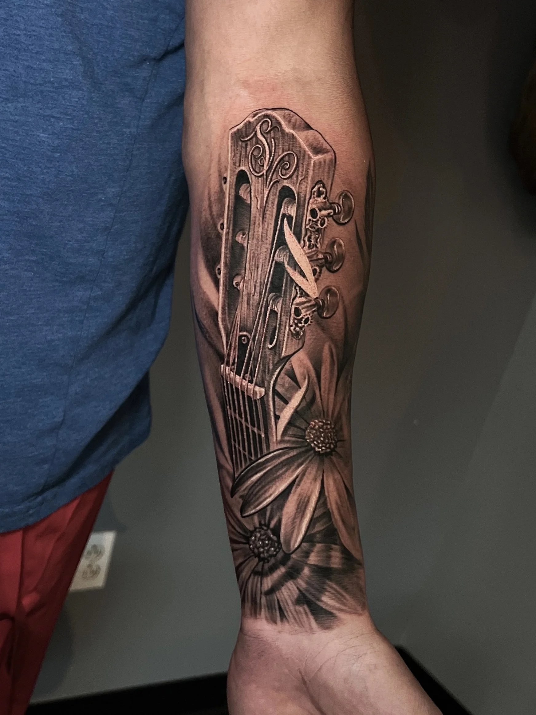Black and gray tattoo of a guitar headstock and flowers on someone's forearm.