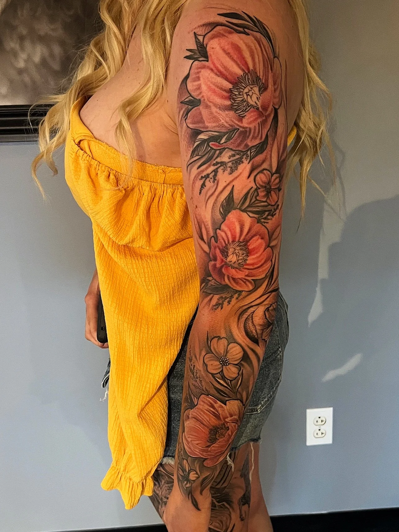 Close-up of a woman's arm with a large floral tattoo featuring orange and pink flowers and green leaves, wearing a yellow sleeveless top.