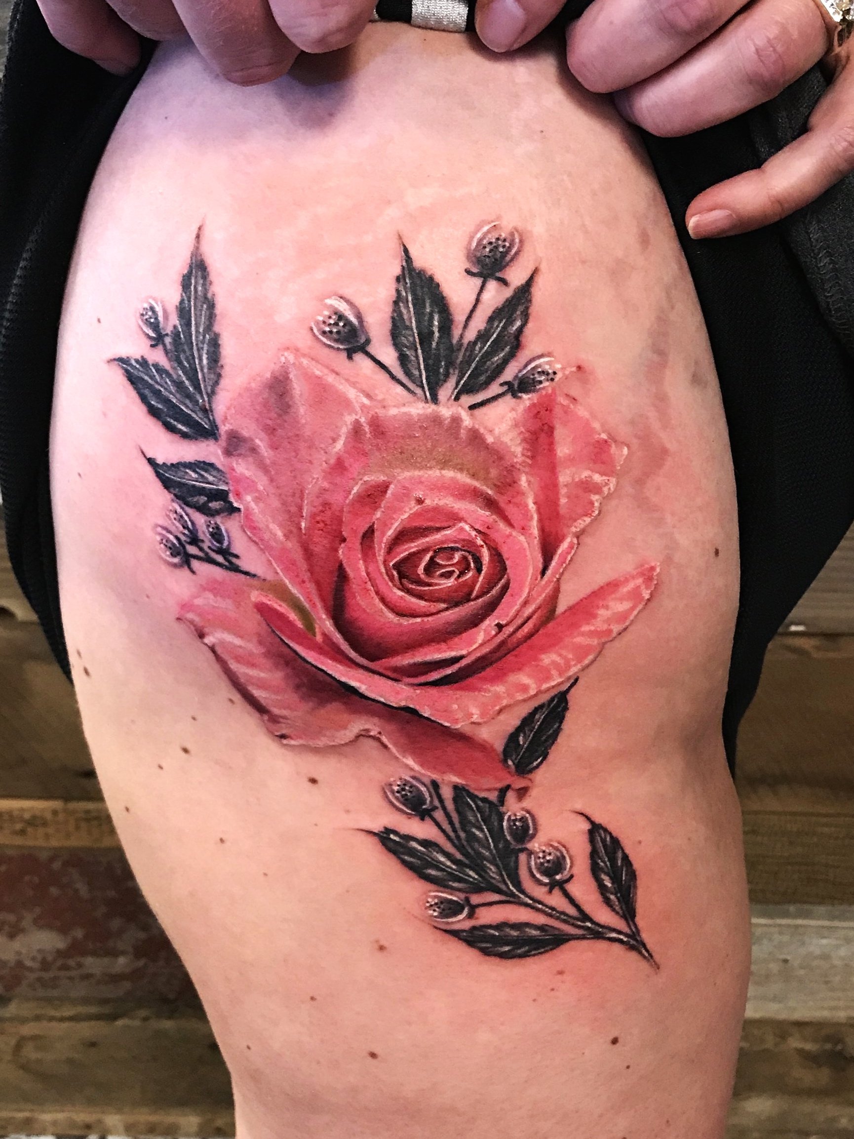 A tattoo of a detailed pink rose with black leaves and small blackberries on the person's upper thigh or hip.