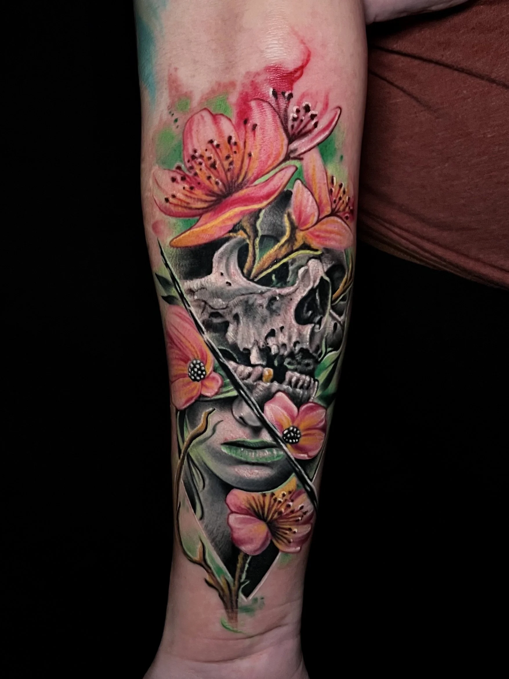 Colorful tattoo on arm featuring pink flowers, a human skull, and a woman's face with closed eyes and green lips.