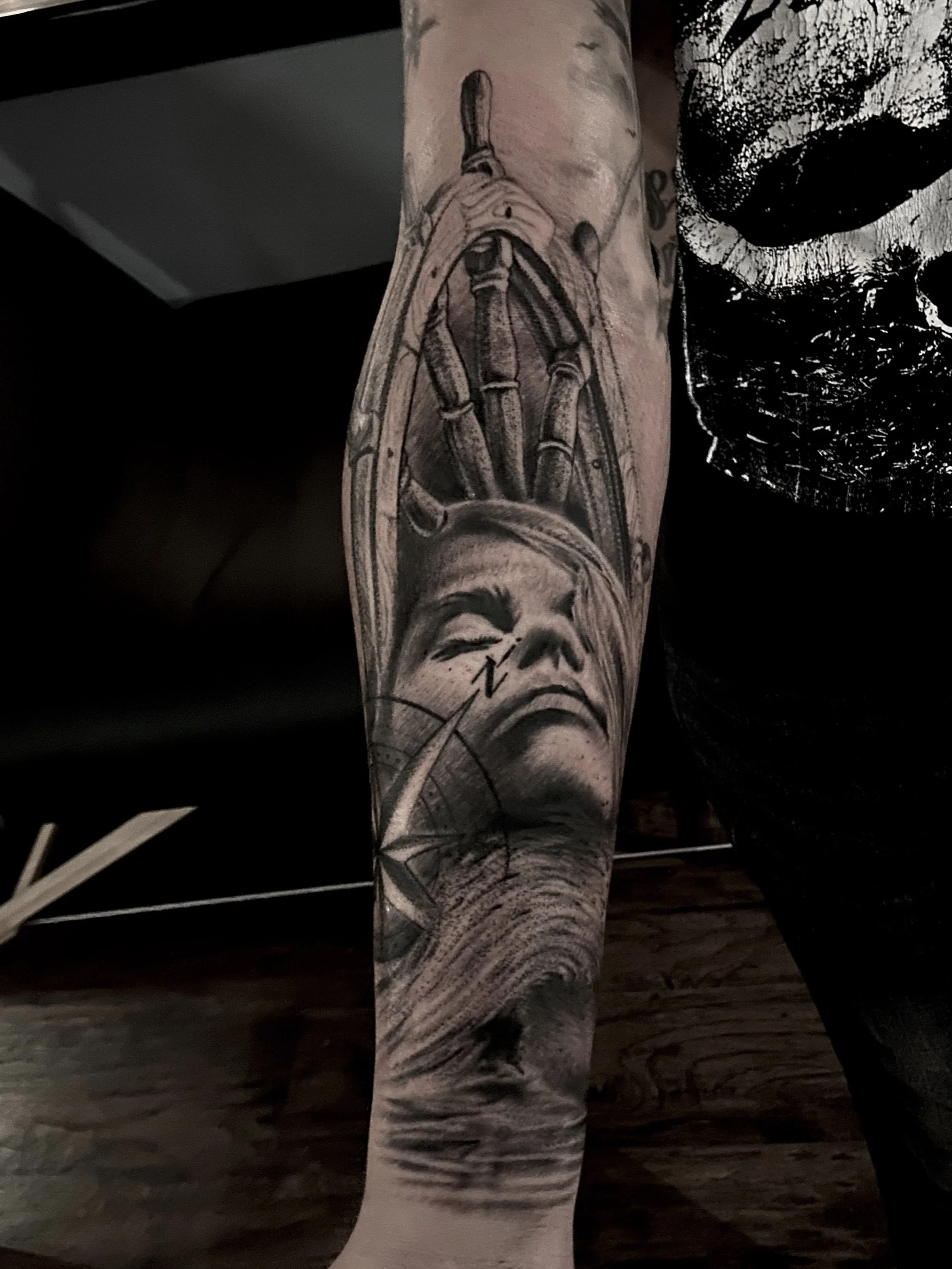 Black and gray tattoo of a woman's face with closed eyes and flowing hair, intersected by a compass, with lighthouse and stormy sea background.