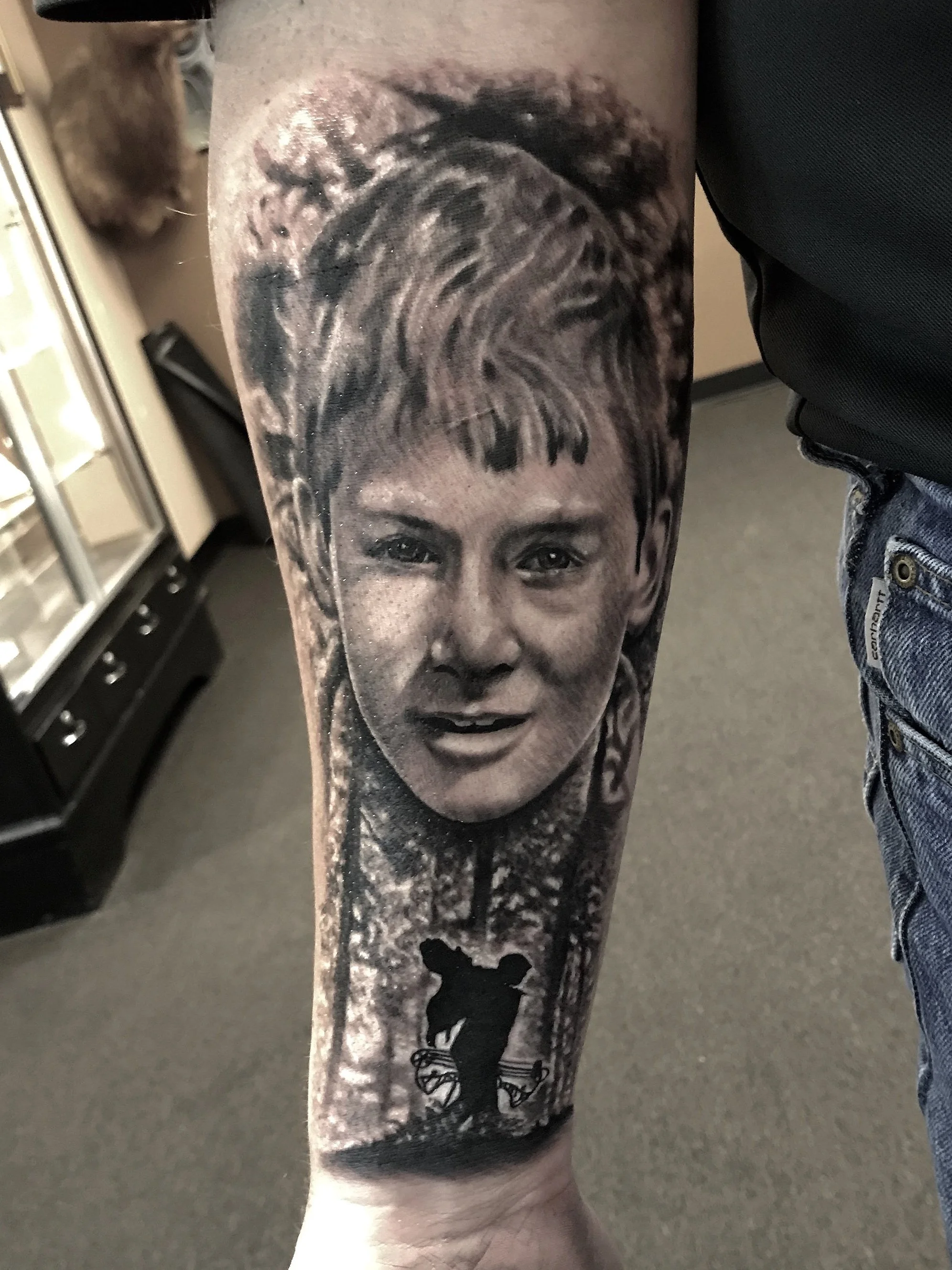Detailed black and gray tattoo on a person's forearm of a young girl's face with short hair, smiling, with trees in the background and a silhouette of two children on a seesaw underneath.