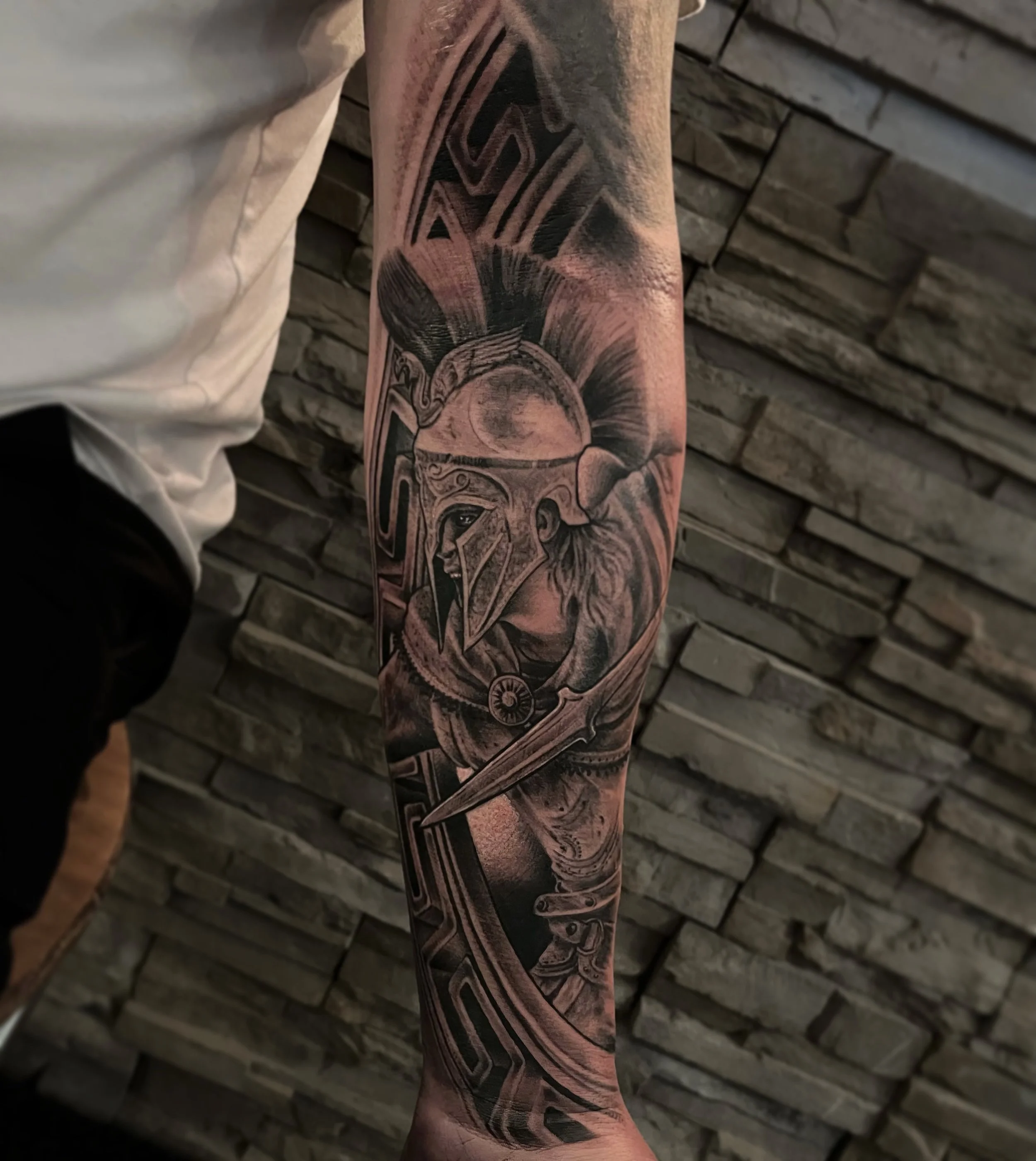 Black and gray tattoo of a warrior wearing a Trojan helmet with a plume, holding a sword, on a person's forearm against a stone wall background.
