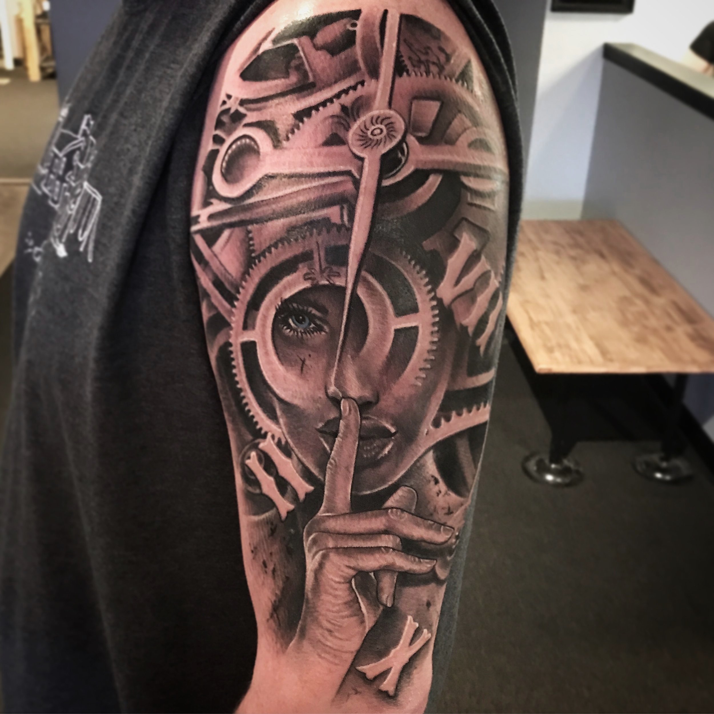 Black and gray tattoo of a woman's face with an eye, surrounded by clock gears and mechanisms, with her finger held to her lips in a shushing gesture.
