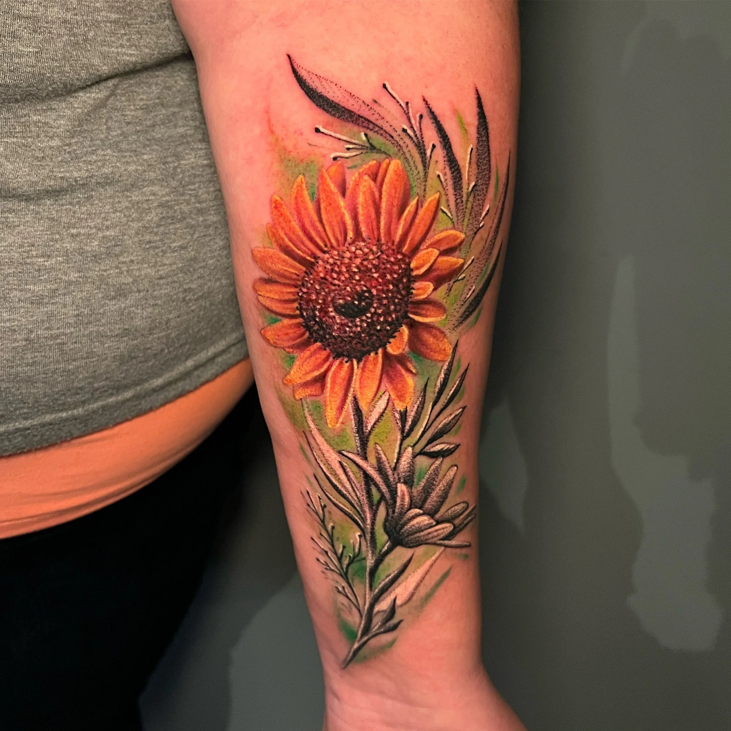 Colorful sunflower tattoo on person's arm with green leaves and black-and-gray outlines.