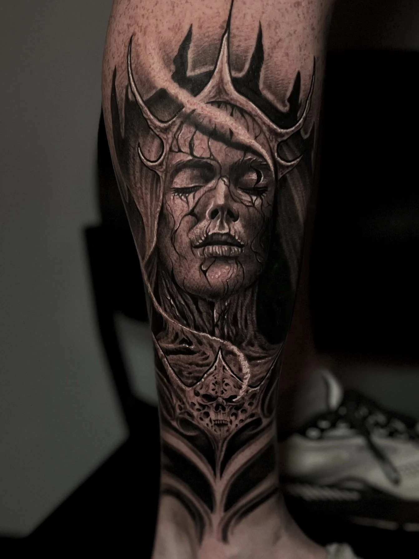 Black and gray tattoo of a female face with closed eyes, cracked skin, and a crown-like structure on her head, featuring a skull at the bottom.