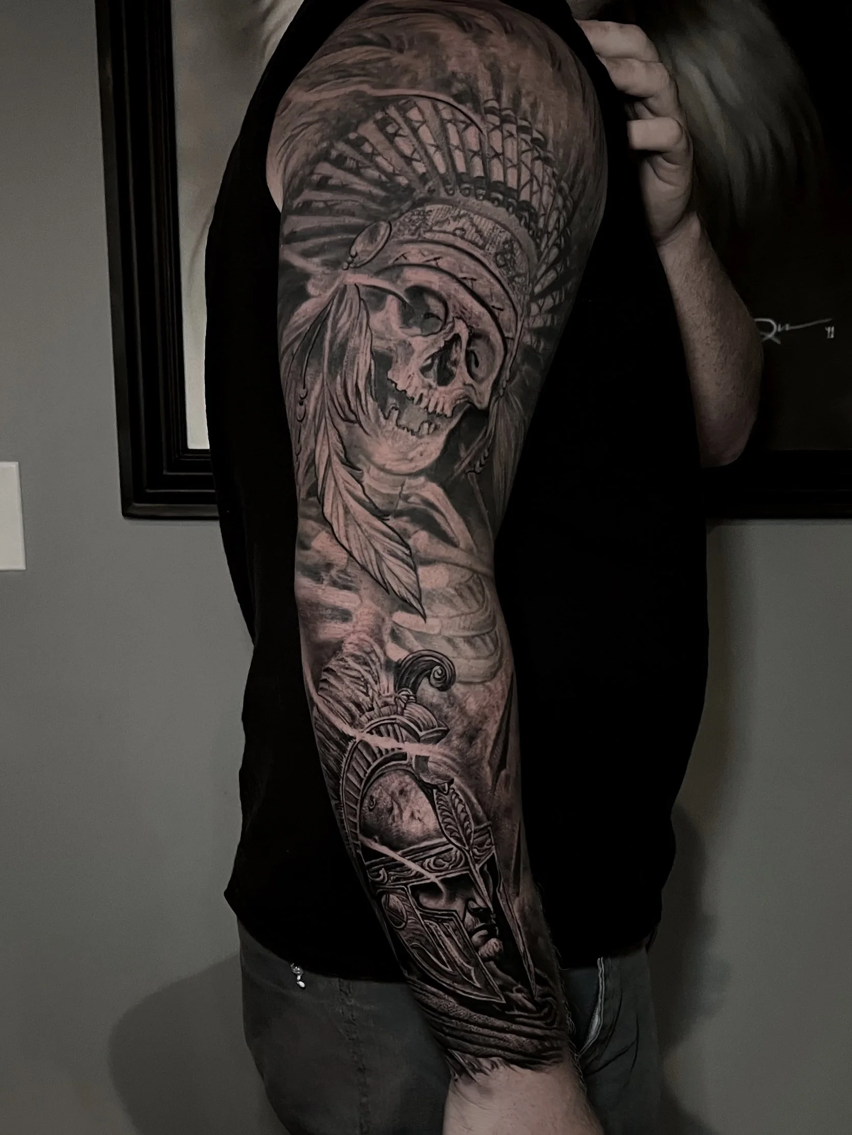 A person with a black sleeveless shirt displaying a detailed black and gray tattoo sleeve. The tattoo features a pirate skull with a pirate hat, feathers, a ship's wheel, and a traditional skull design, covering the upper arm and forearm.