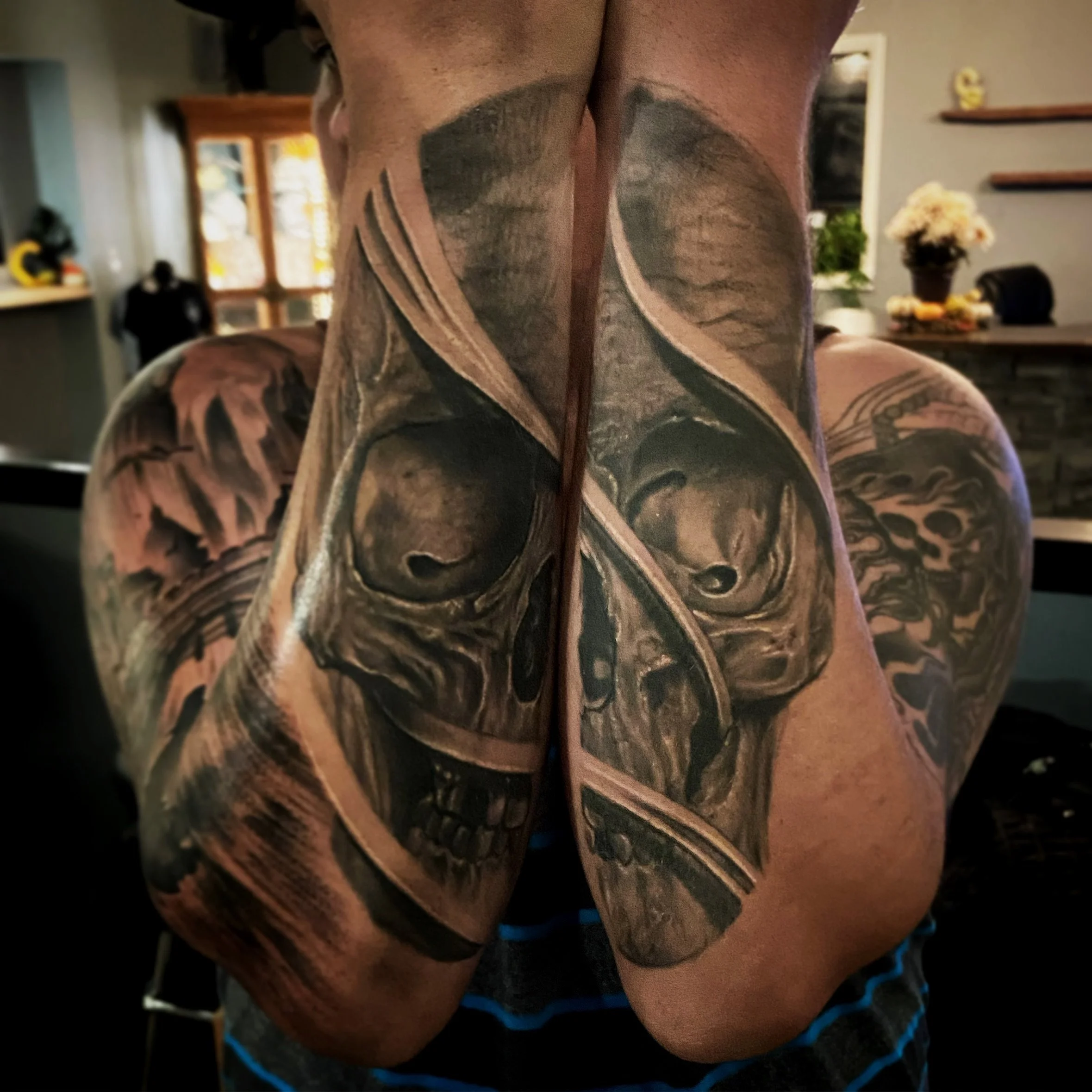 A person with detailed black and gray tattoo of skulls and abstract designs on both arms in a living room setting.