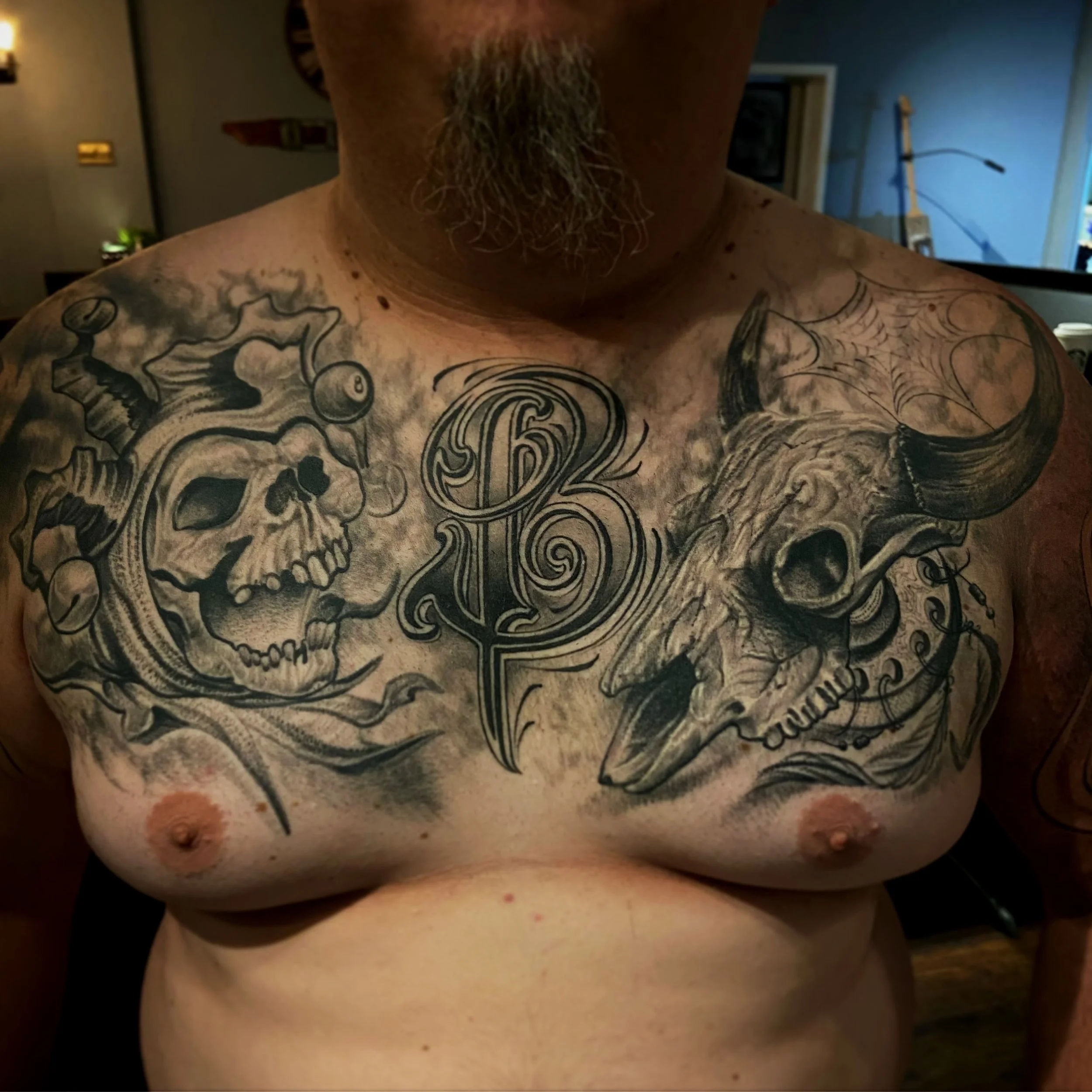 Tattooed man's chest featuring a skull with a mermaid tail, a stylized letter B, and a skull with horns surrounded by a spider web.