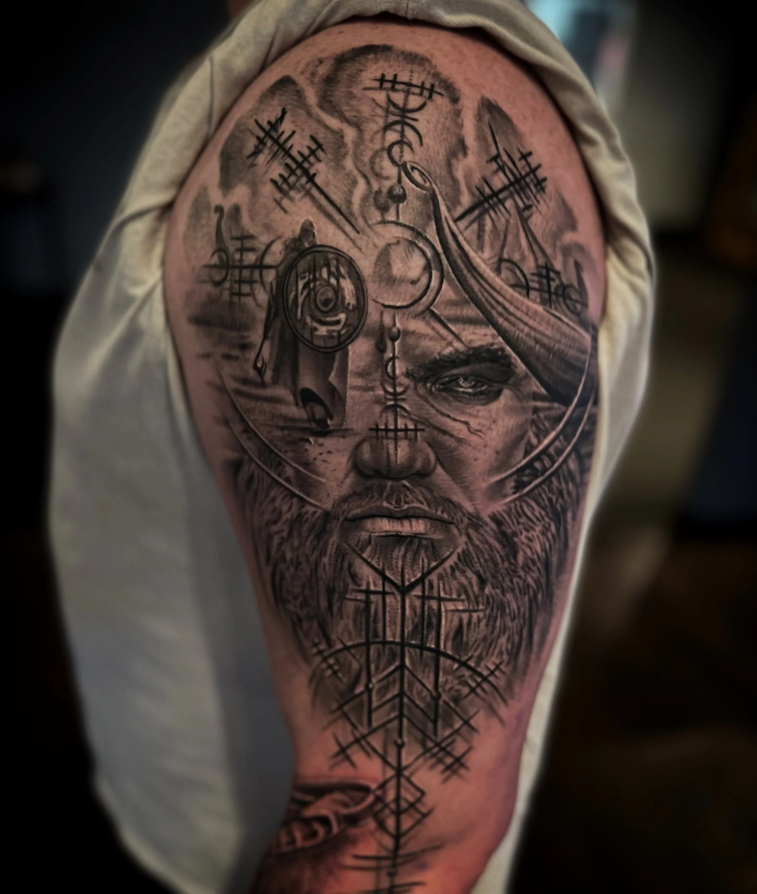 Black and gray tattoo of a mystical face with symbols, shapes, and a beard on a person's upper arm.