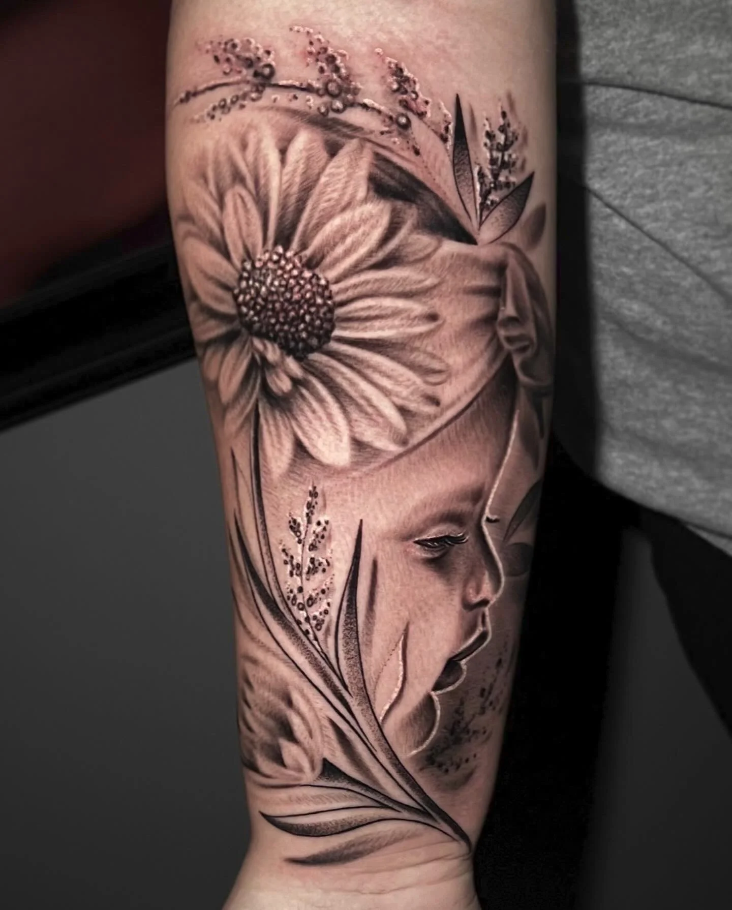 Black and gray tattoo of a girl's face in profile with a large flower above her head and decorative leaves and smaller flowers surrounding her.