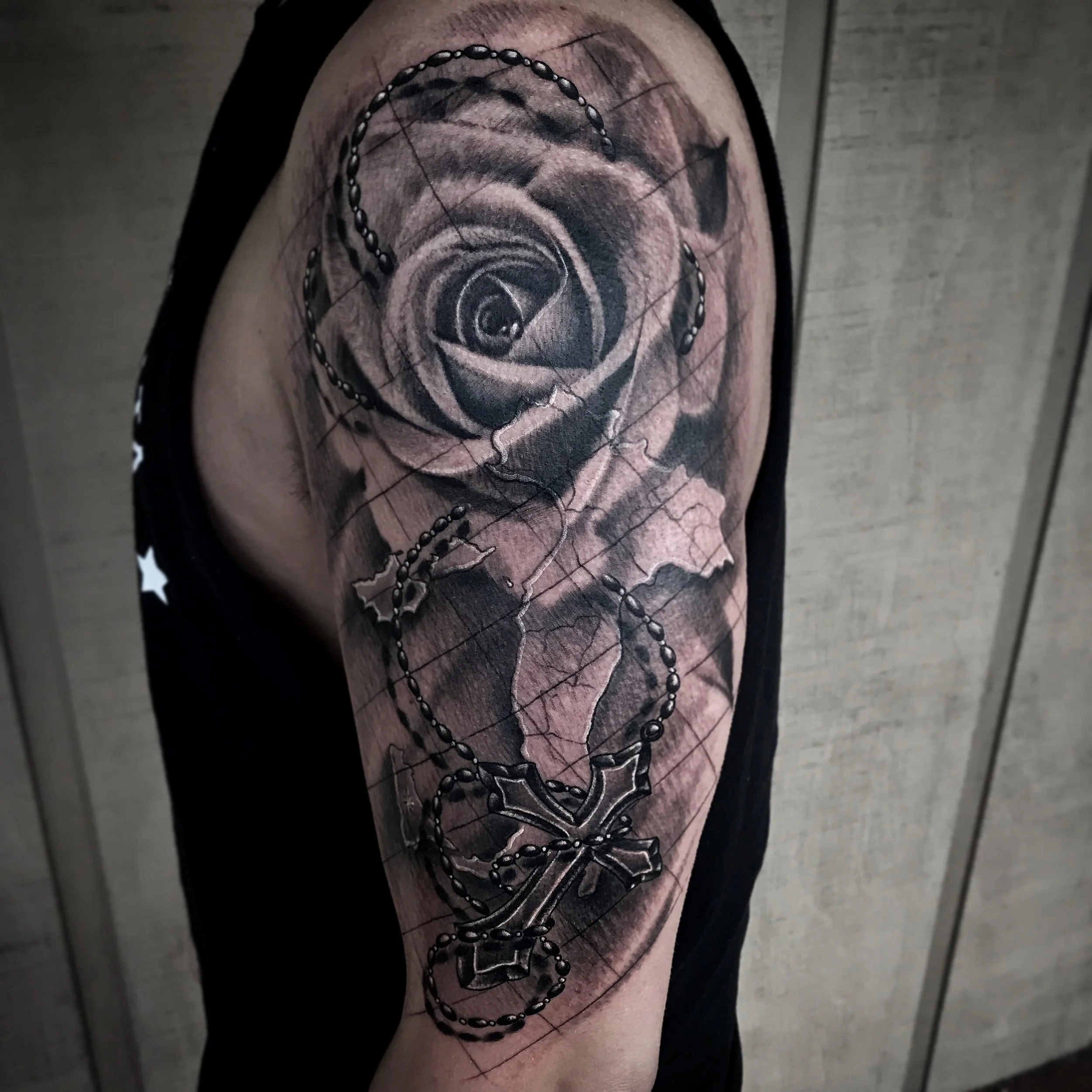 Black and gray tattoo on person's upper arm featuring a detailed rose, a map of Alabama, and a rosary with a cross pendant.