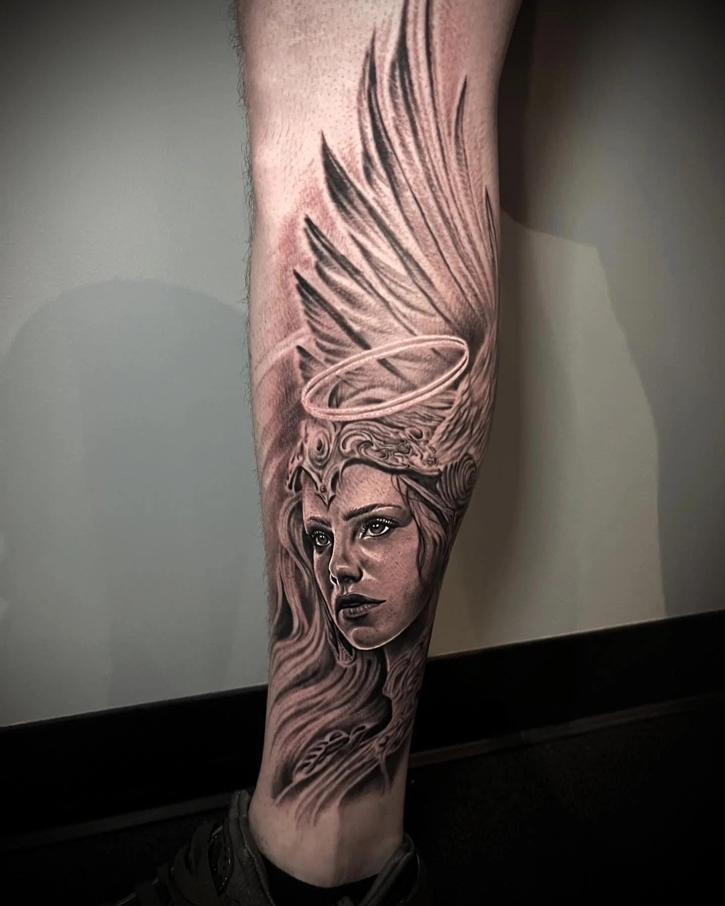 Black and gray realism tattoo of a woman with flowing hair, angel wings, and a halo on a person's calf.