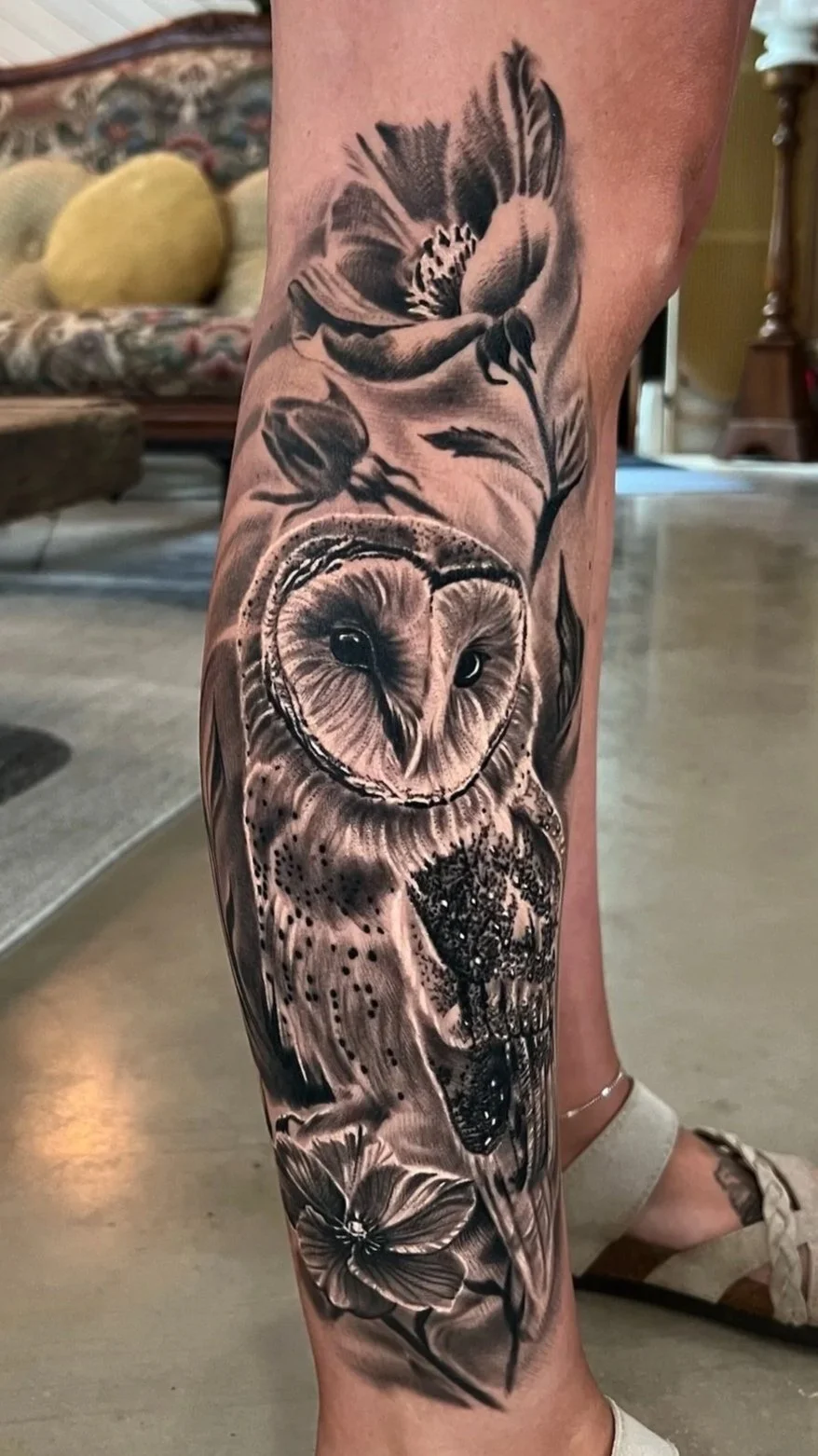 Black and gray tattoo of an owl with a large flower and leaves on a person's lower leg.