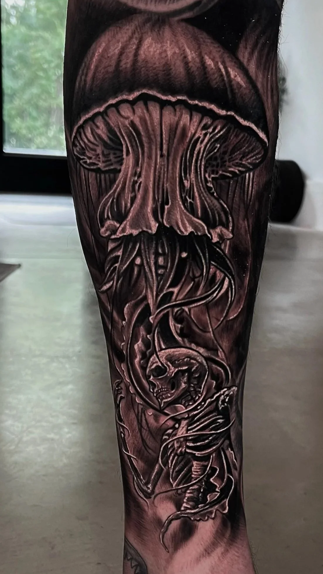 Black and gray tattoo of a mushroom with a gilled cap, beneath a skull with a snake wrapped around its jaw, on a person's arm.