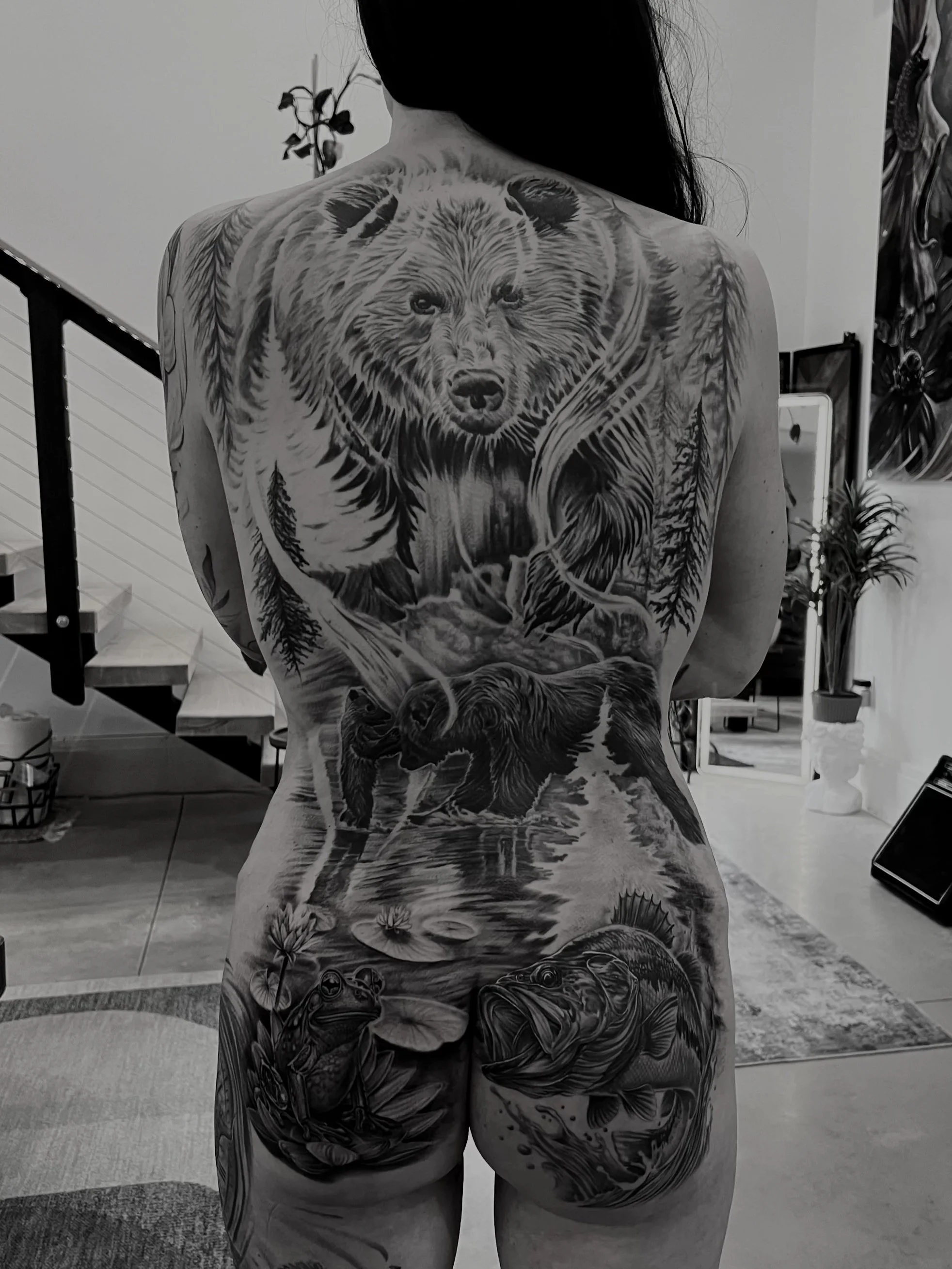 Back of person with detailed black and gray tattoo of a bear, wolves, fish, and nature scene covering their back and buttocks.