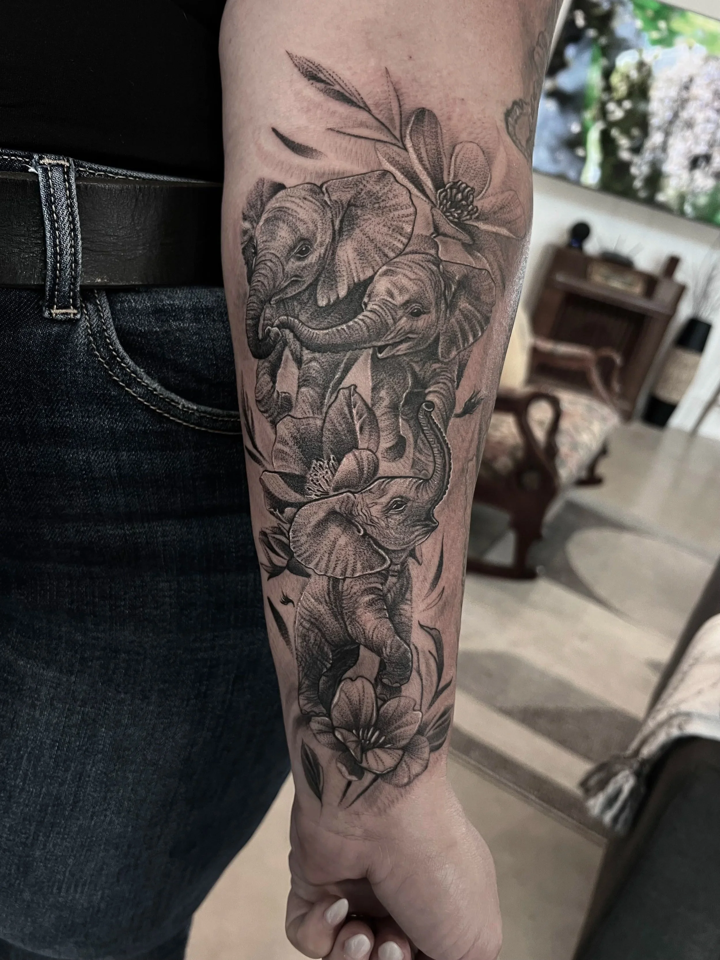 A detailed black and gray tattoo of three elephants surrounded by flowers on a person's forearm.