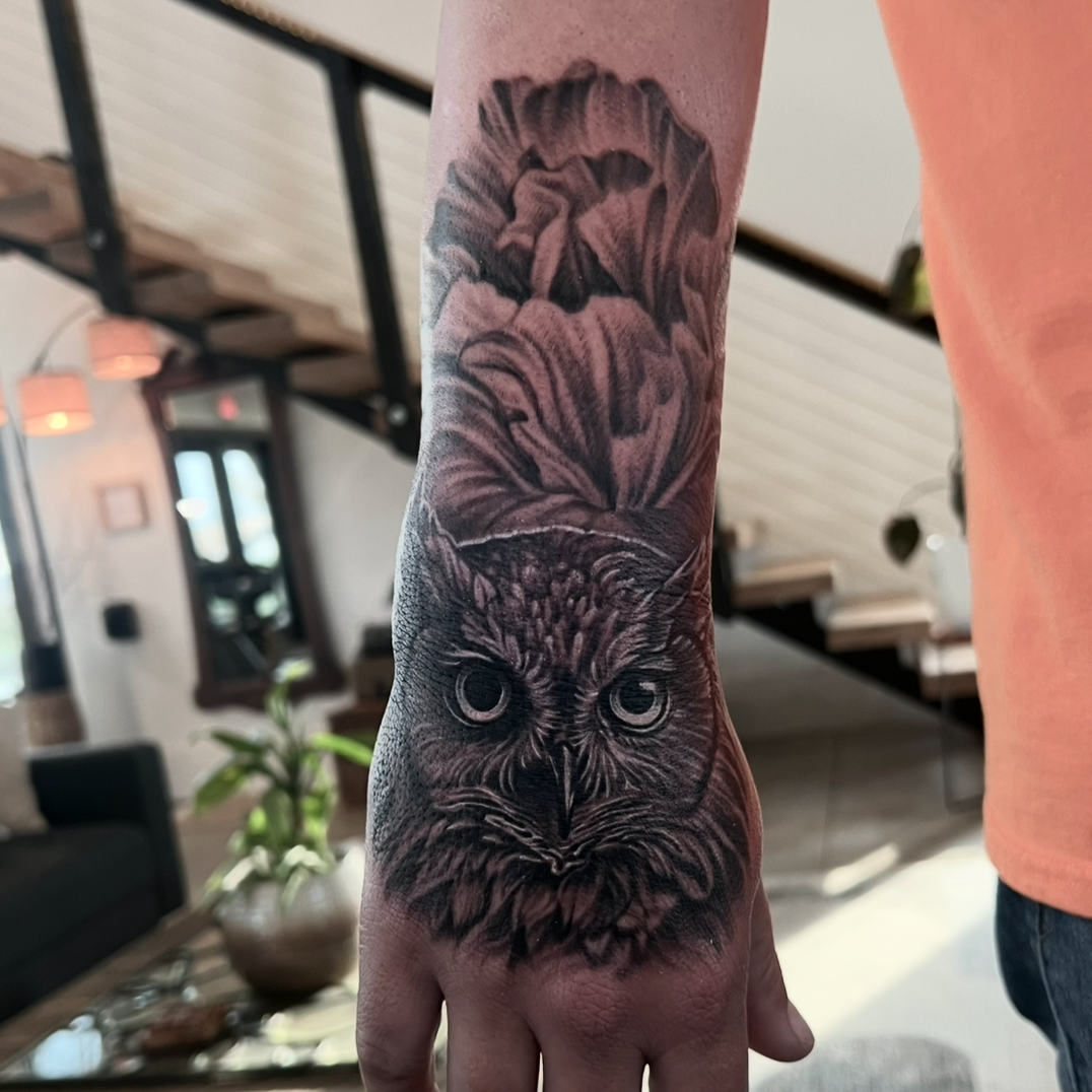 Black and gray tattoo of an owl's face on a person's forearm with detailed feathers and intense eyes.
