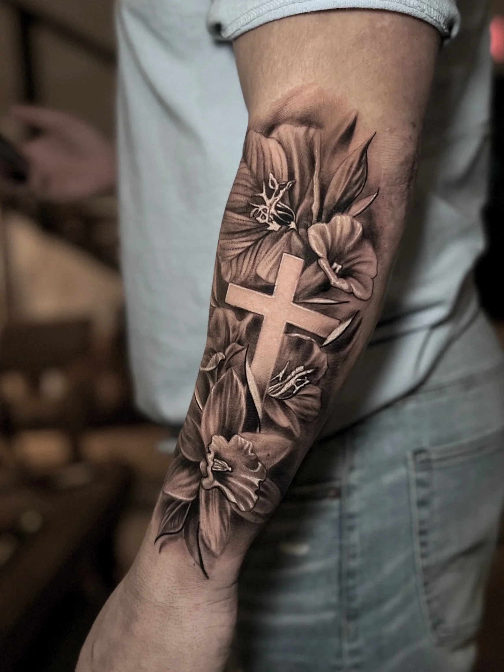 Black and gray tattoo on person's forearm depicting a cross surrounded by large, detailed lilies and leaves.