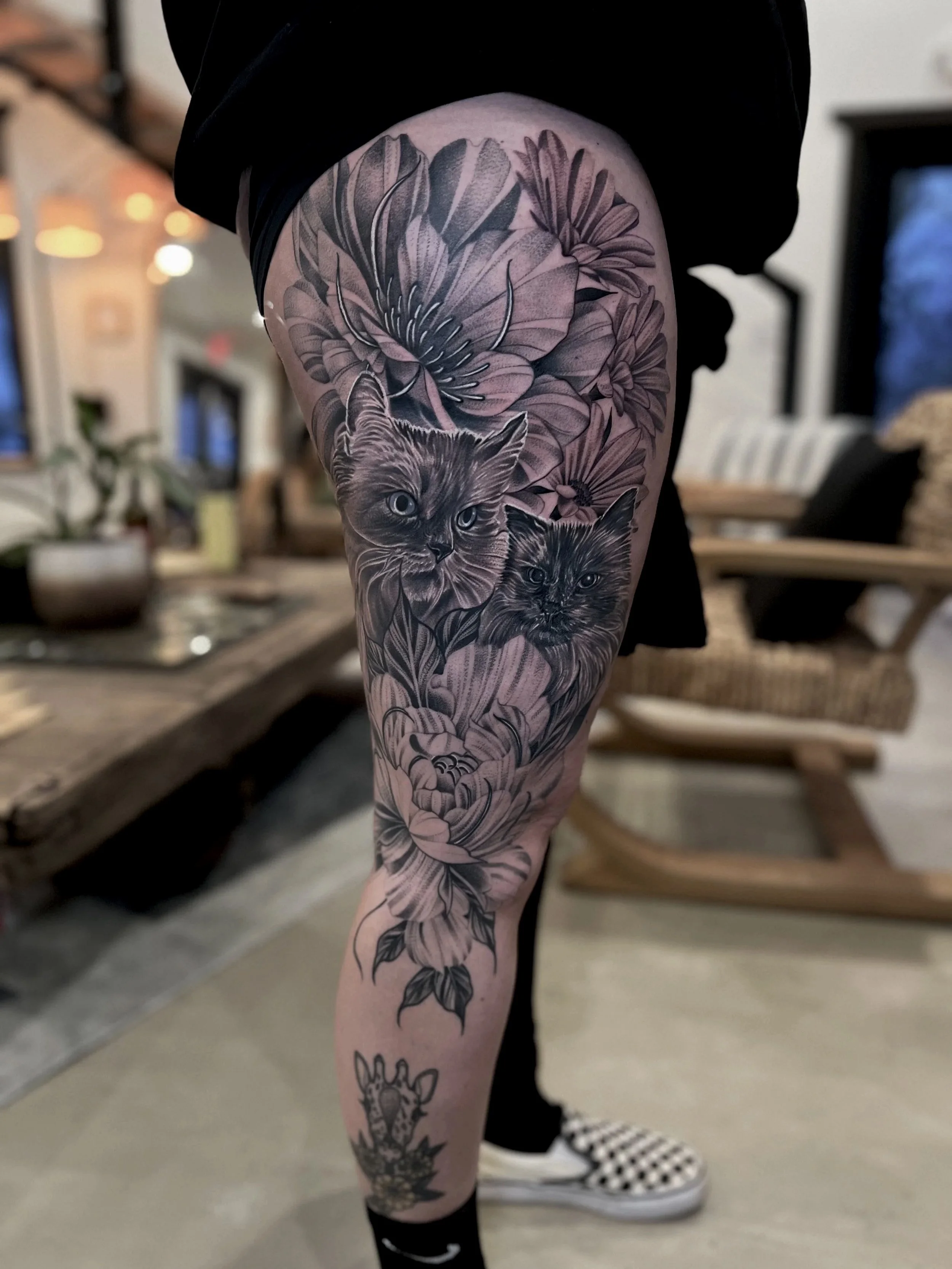 Black and gray tattoo sleeve featuring two detailed cat faces and large flowers on person's thigh.